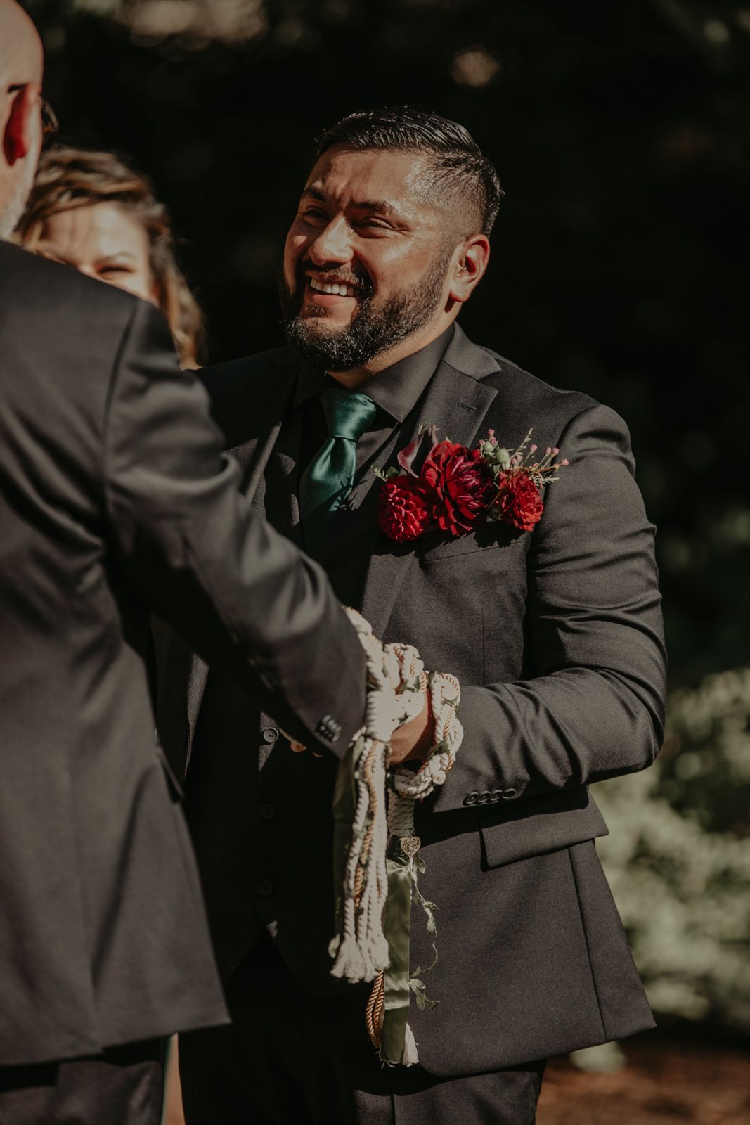 A groomsman in a dark suit with red boutonniere laughs joyfully during an outdoor wedding celebration.