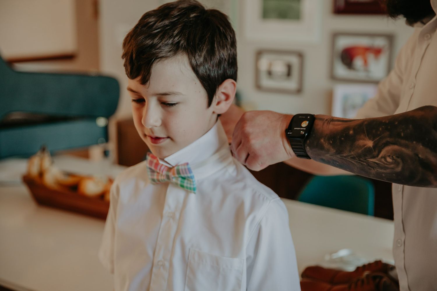 Someone wearing a white dress shirt has a floral bow tie adjusted by a tattooed hand in an indoor setting.