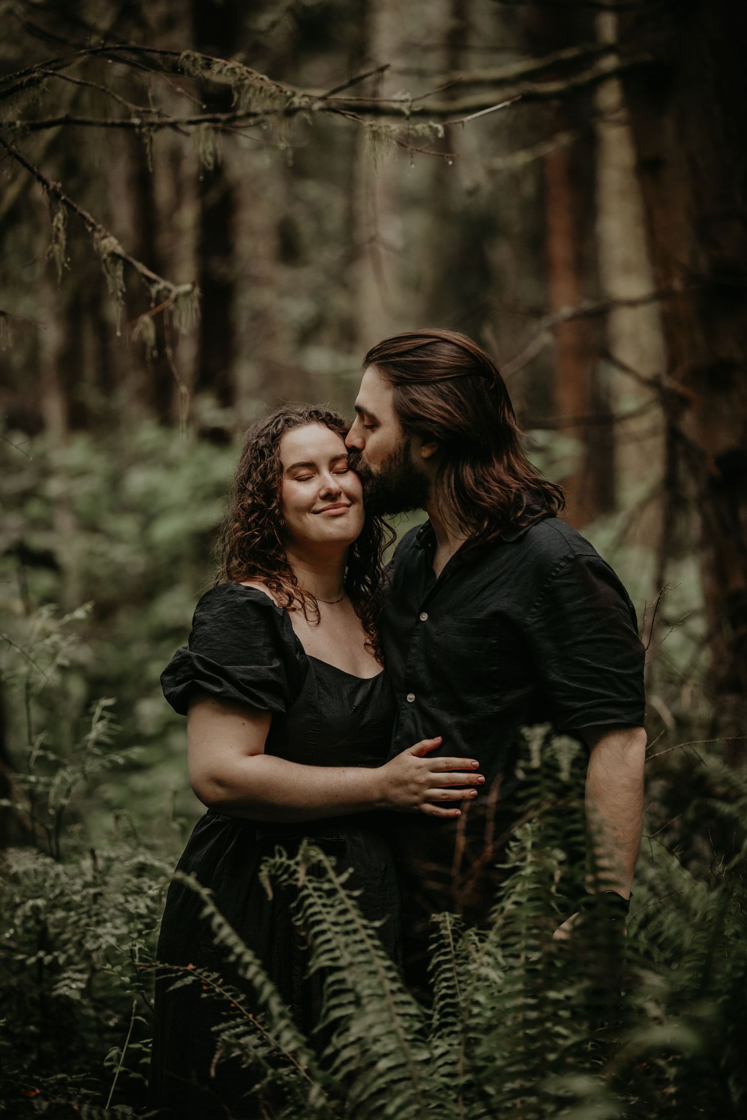 A loving couple embraces in a moody forest setting surrounded by ferns and trees.