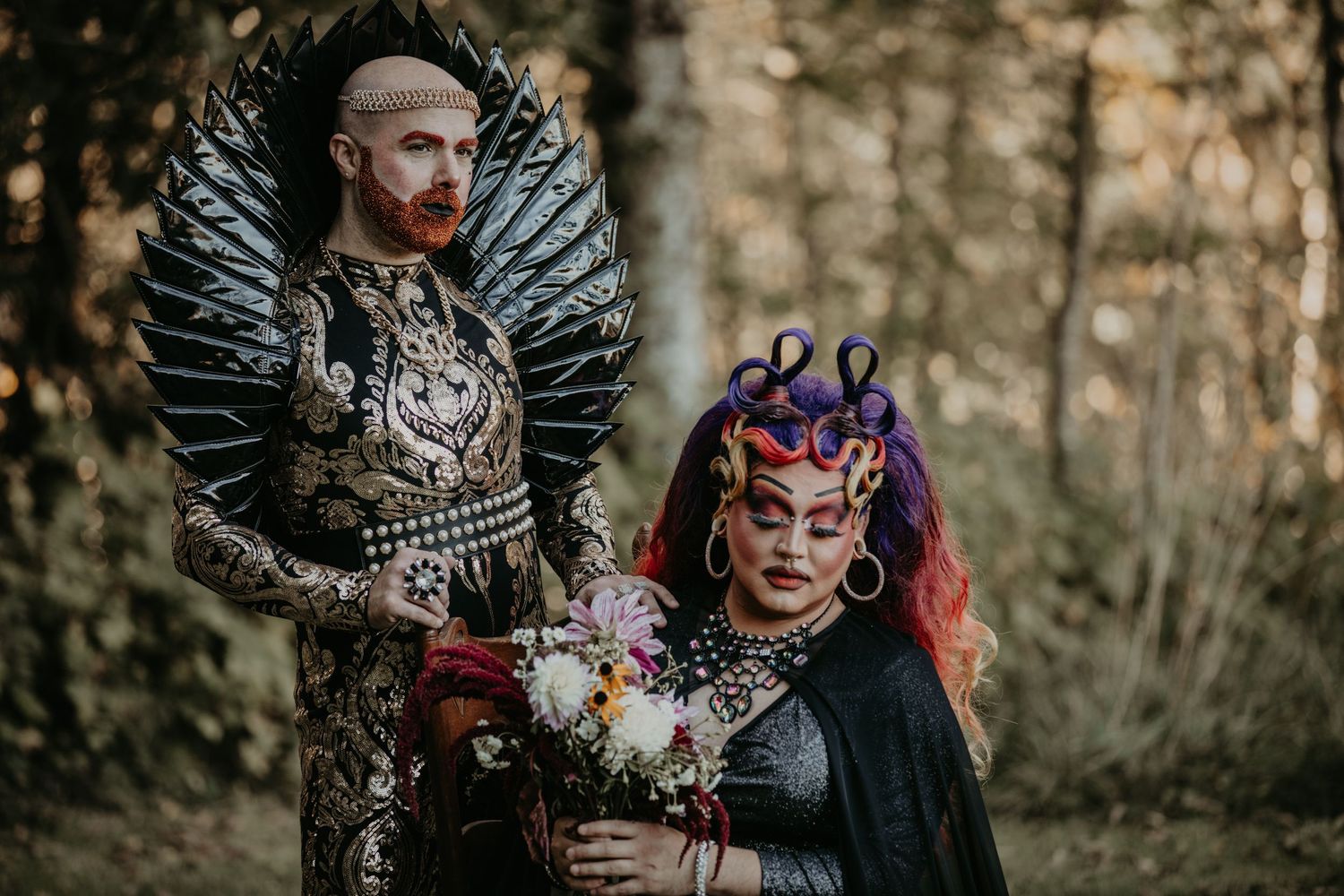 Two figures in elaborate dark fantasy costumes featuring wings and ornate details pose together in an outdoor setting.