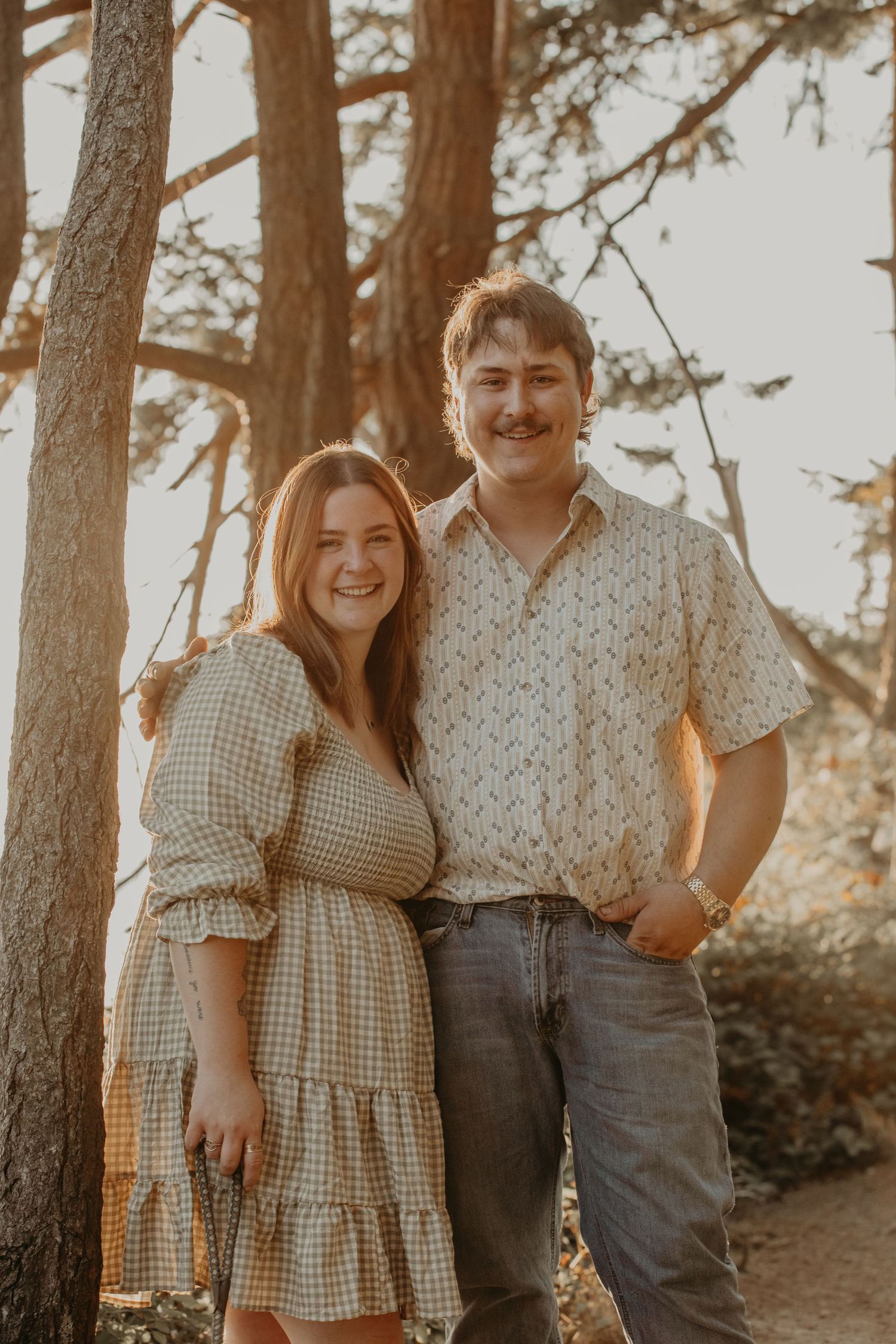 A couple laughs together during a casual outdoor photoshoot among pine trees at sunset.