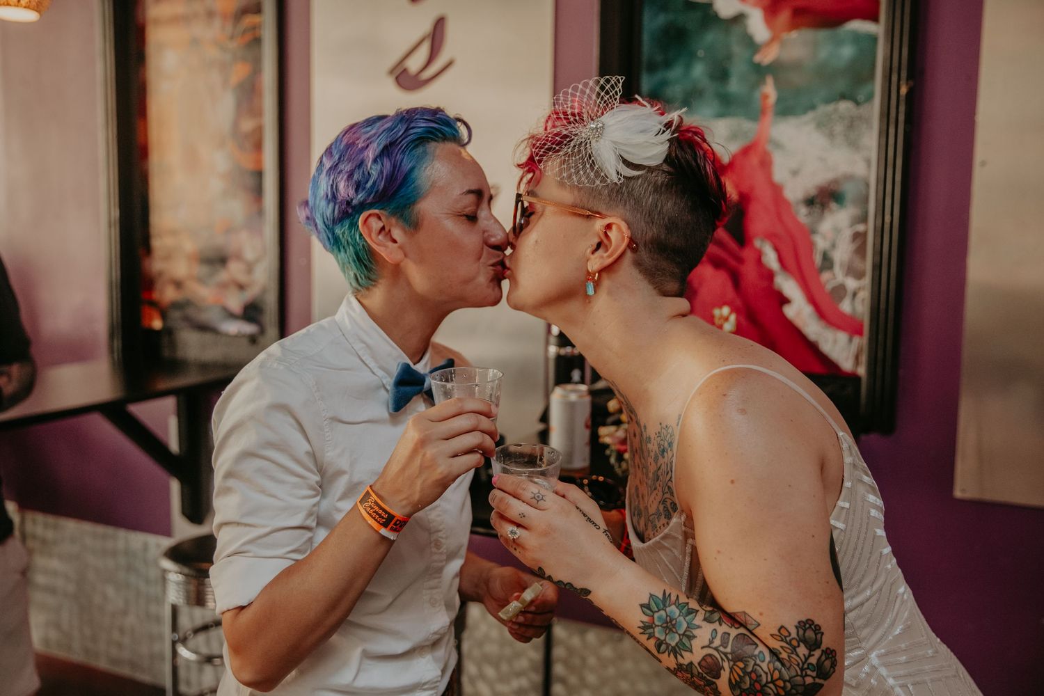 Two people share a romantic kiss while holding drinks at a colorful bar setting.