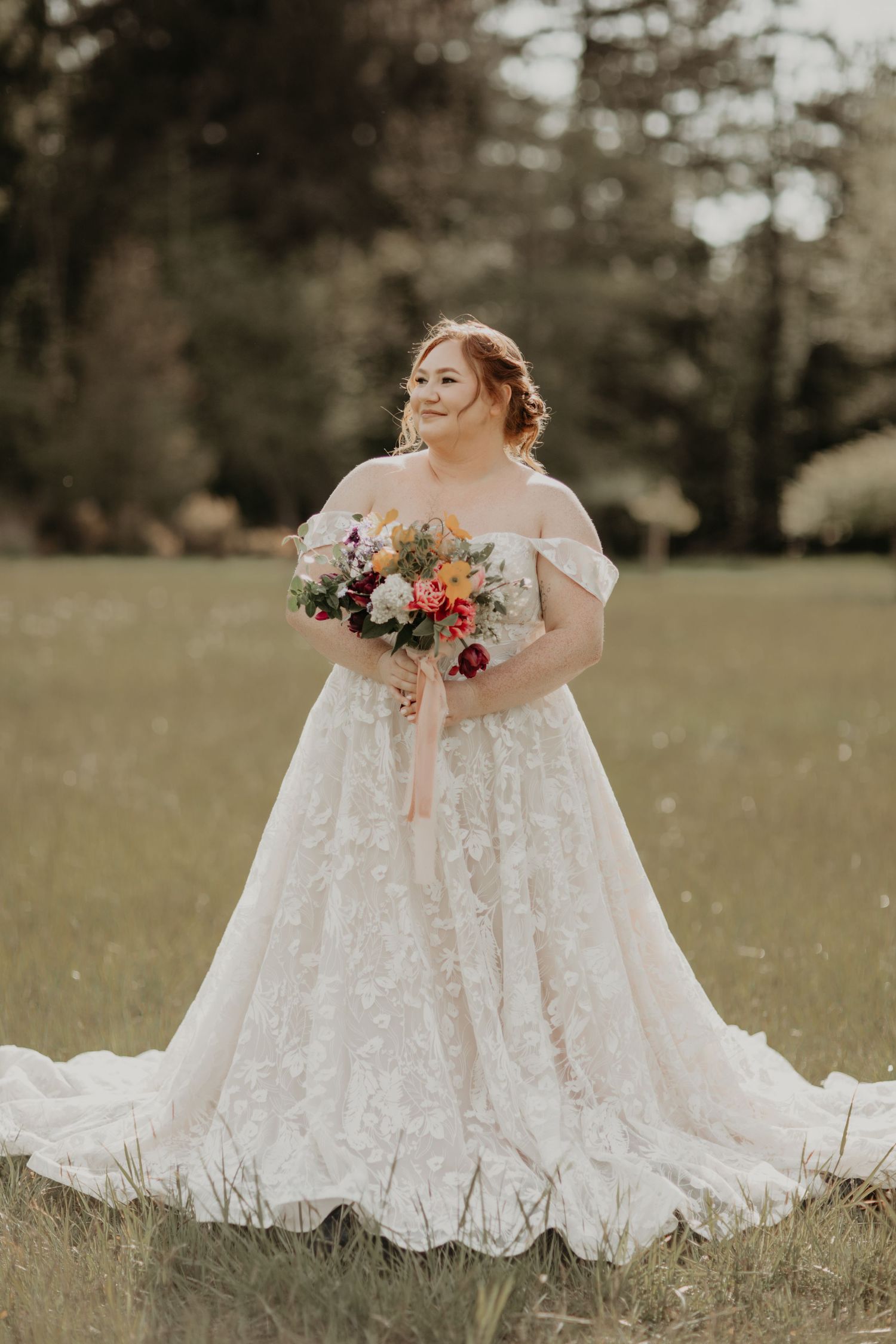 Bride in lace wedding dress holds colorful bouquet while sitting in grassy field during outdoor photo session.