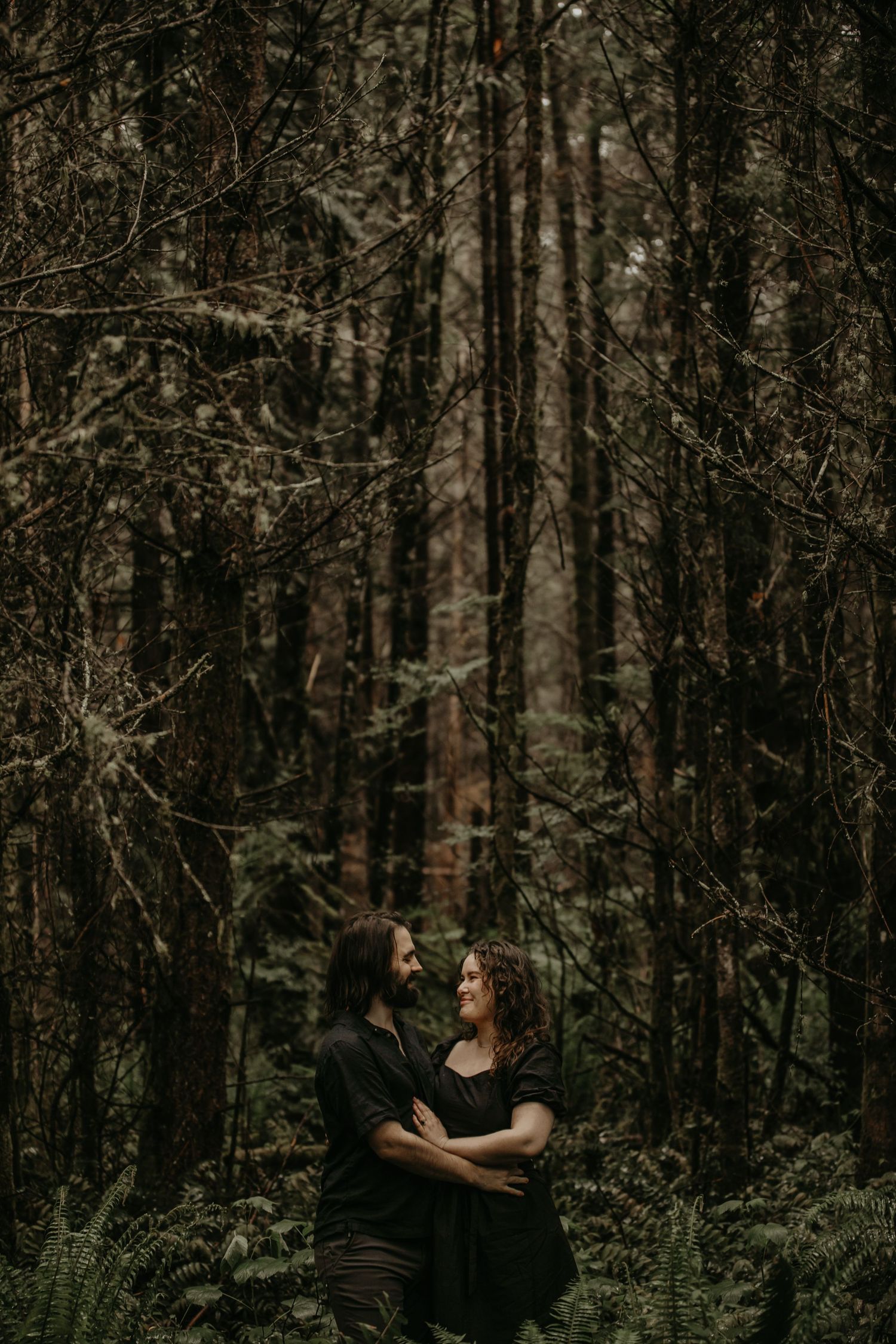 A romantic couple embraces in a moody forest setting with tall trees and ferns creating a mystical atmosphere.