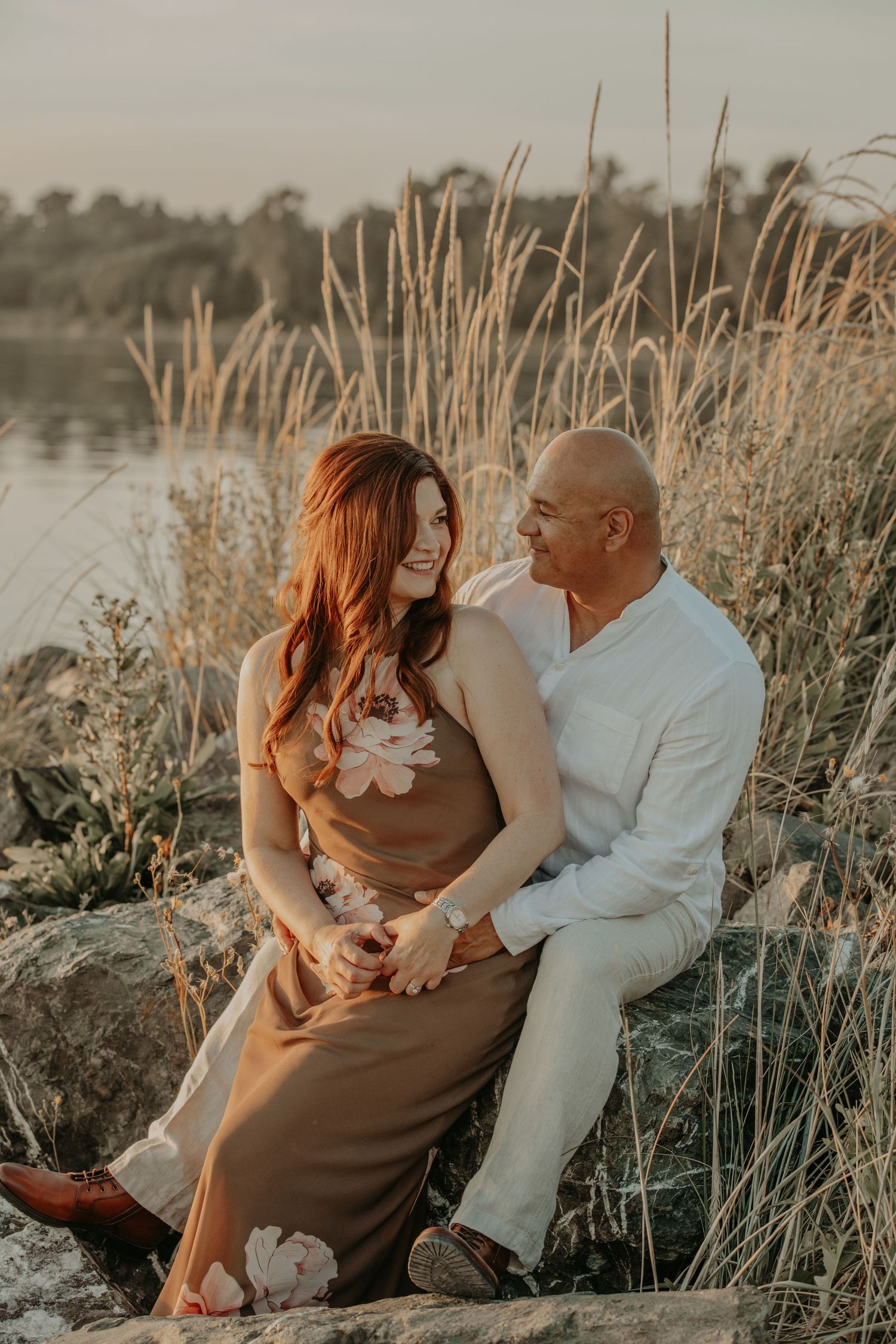 A couple sits together on rocks by a lake at sunset, surrounded by tall golden grasses and warm light.