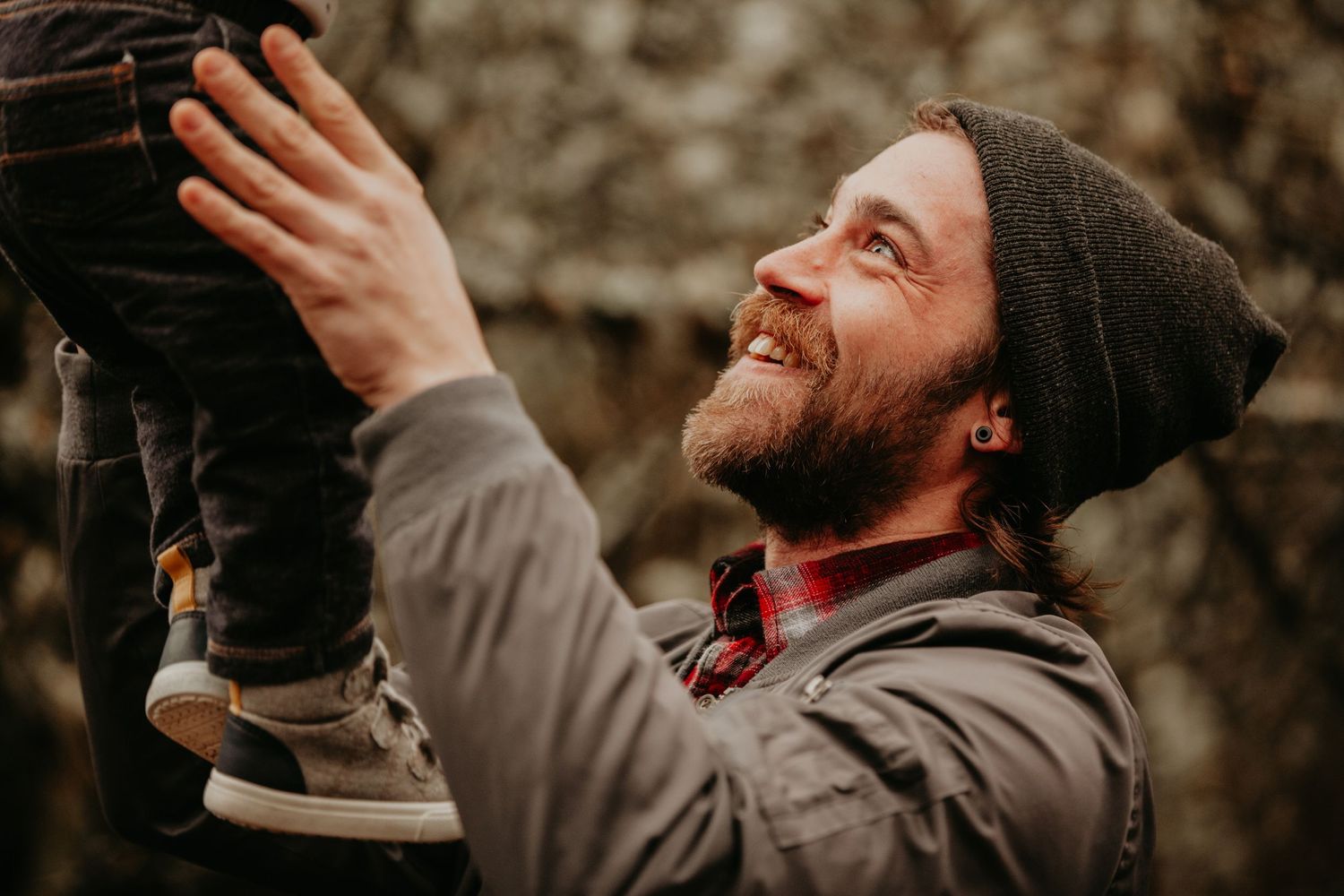 Bearded person in beanie and jacket smiling while looking up and touching hanging lights outdoors.