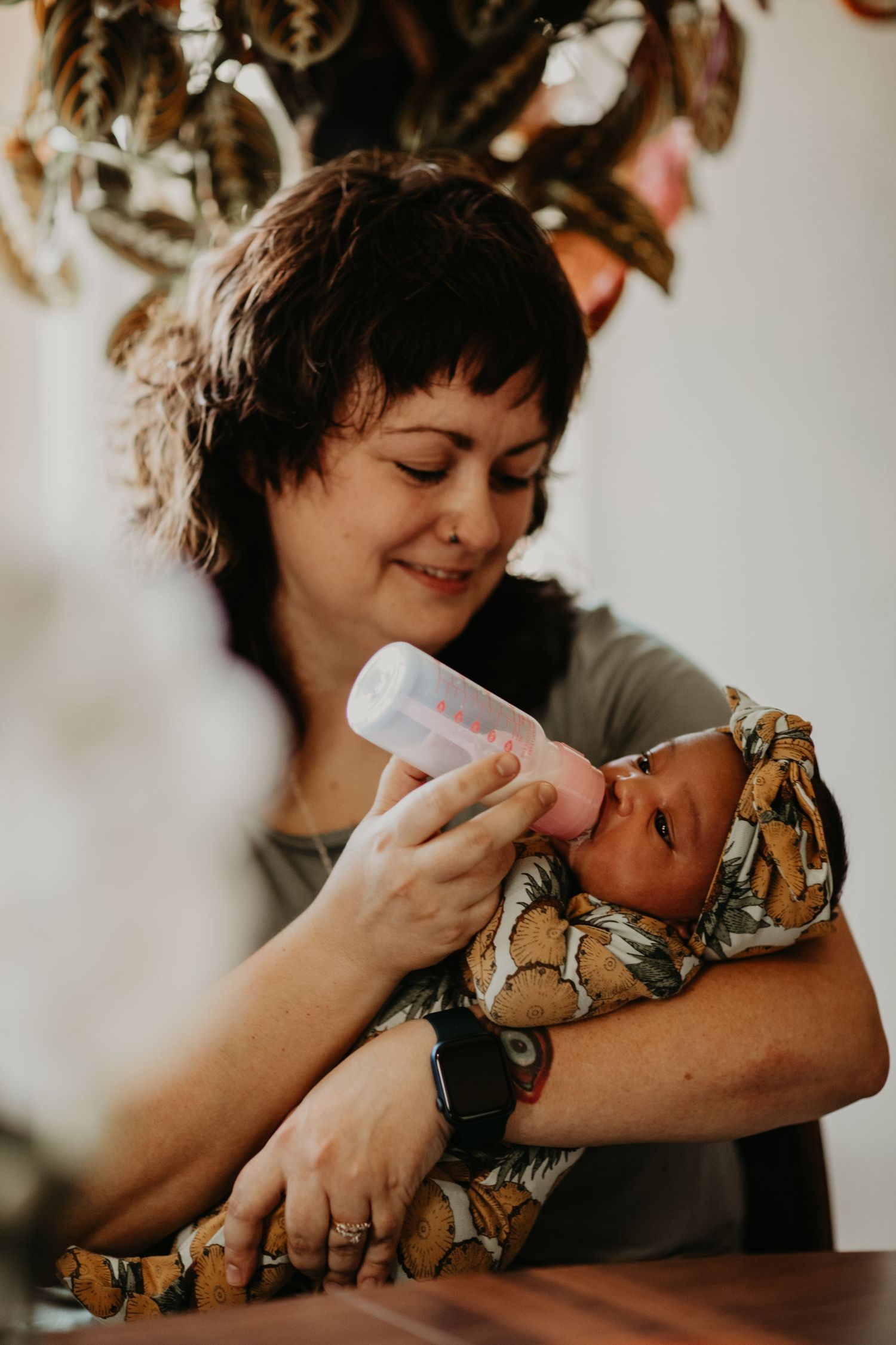 A heartwarming moment as a parent bottle feeds their newborn baby while sitting at a wooden table under indoor plants.