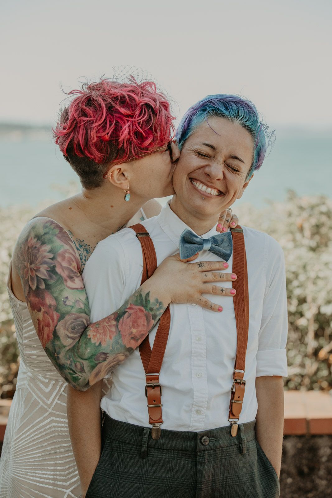 A joyful moment between a couple wearing suspenders and a white dress against an ocean backdrop.