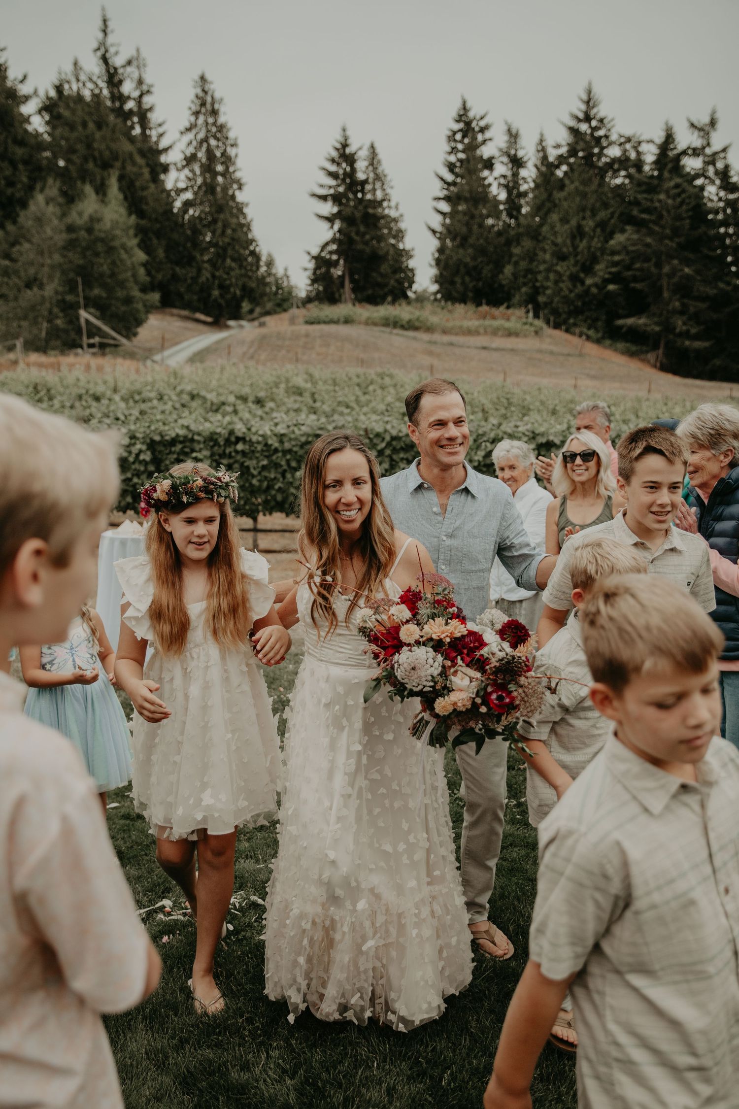 A joyful group in light colored attire dances together in a grassy outdoor setting with evergreen trees in the background.