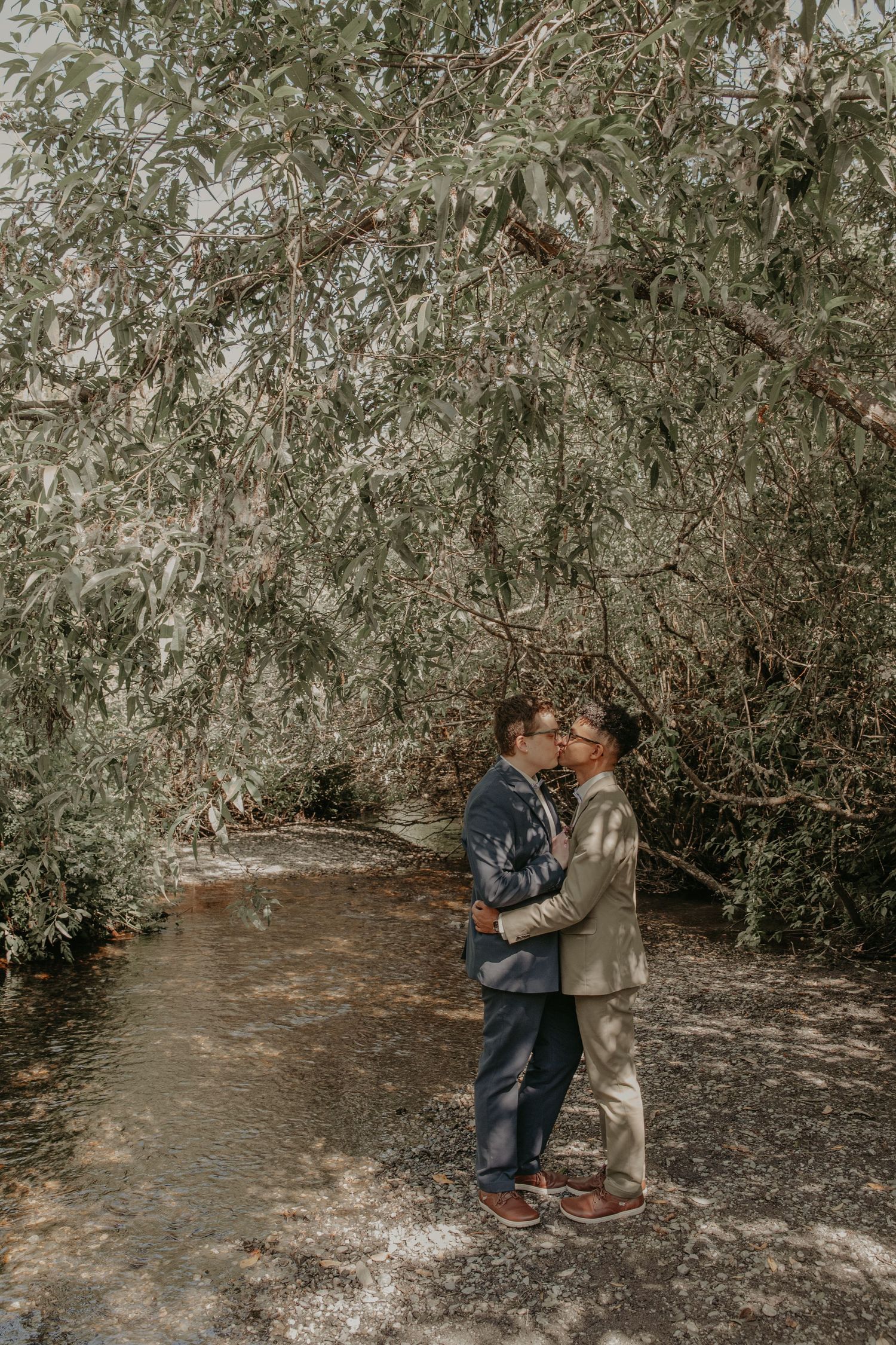 A romantic couple embraces on a path surrounded by olive trees on an overcast day.