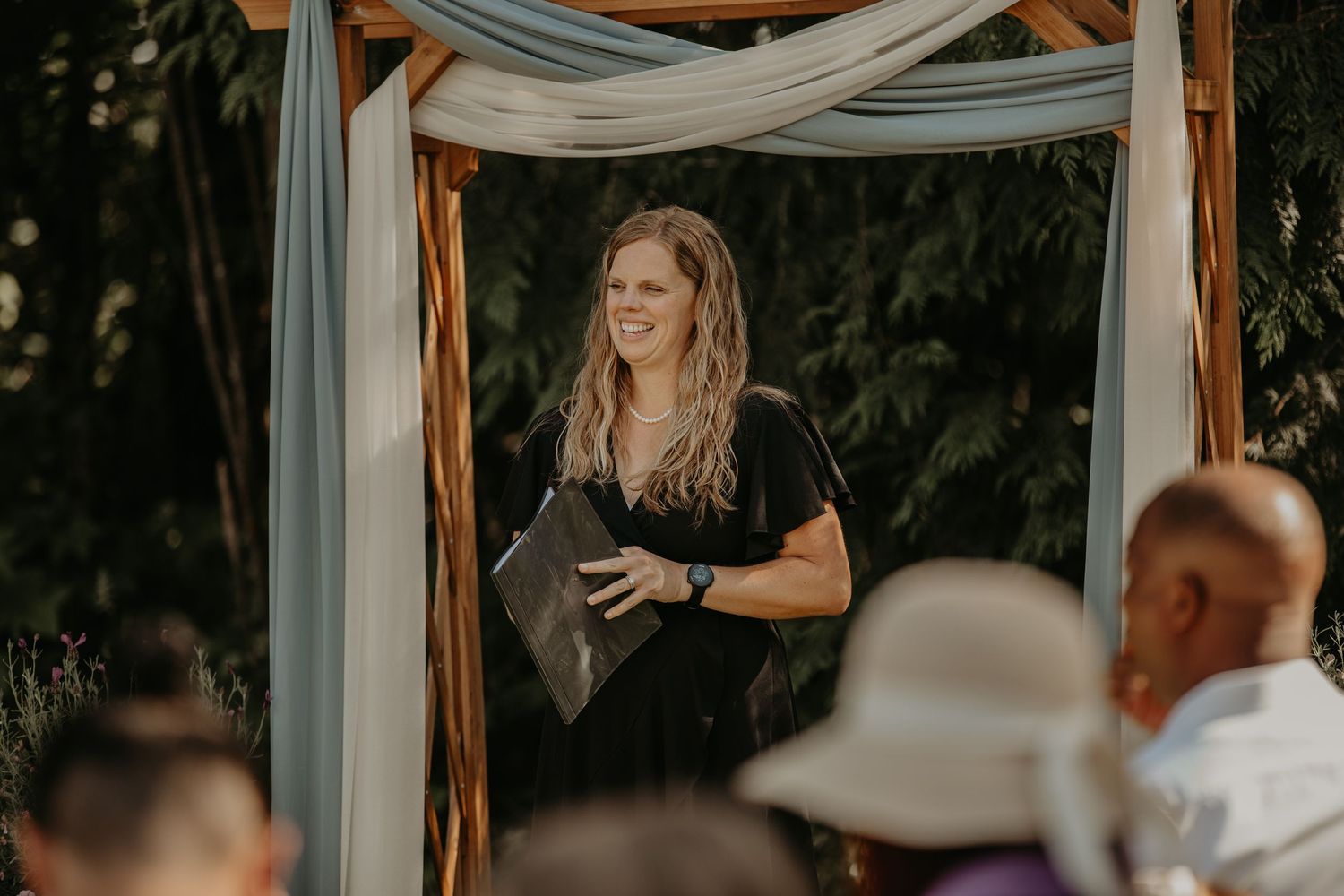 Rustic outdoor wedding ceremony space with white fabric draping and wooden beams against a wooded backdrop.