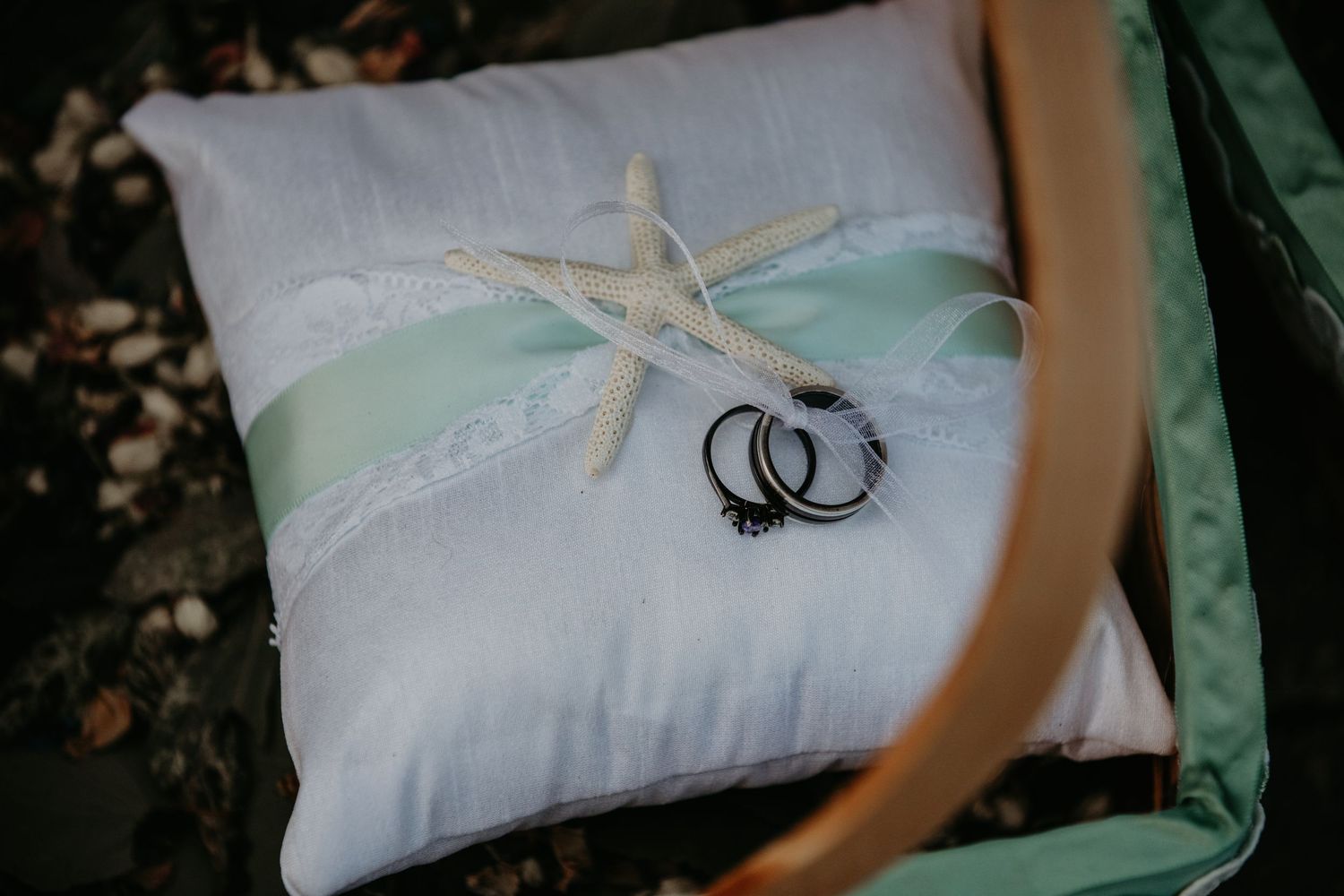 White wedding ring pillow decorated with mint green ribbon, lace trim, starfish accent and two silver wedding bands.