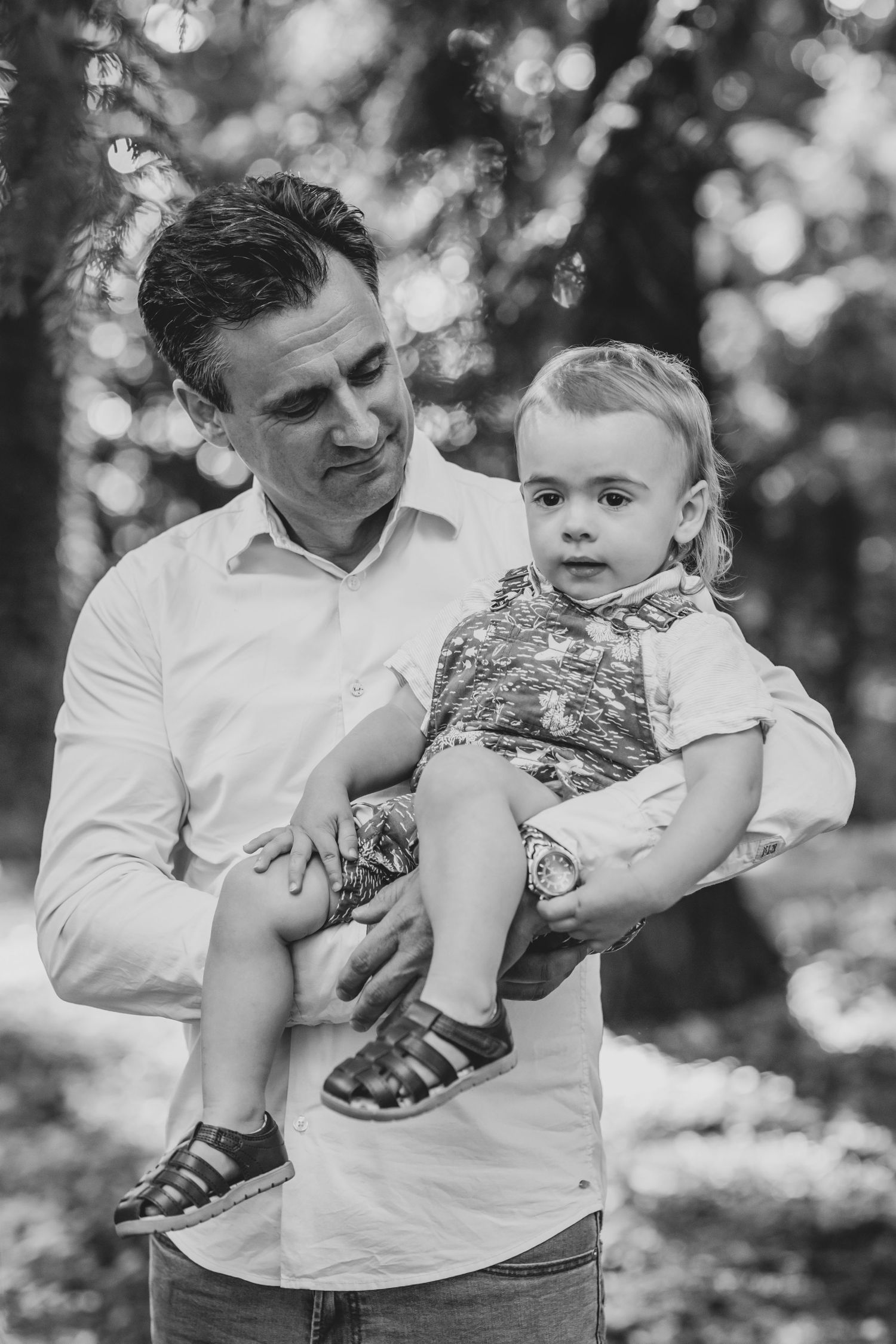 A black and white portrait of a parent holding a small toddler outdoors with a soft bokeh background.