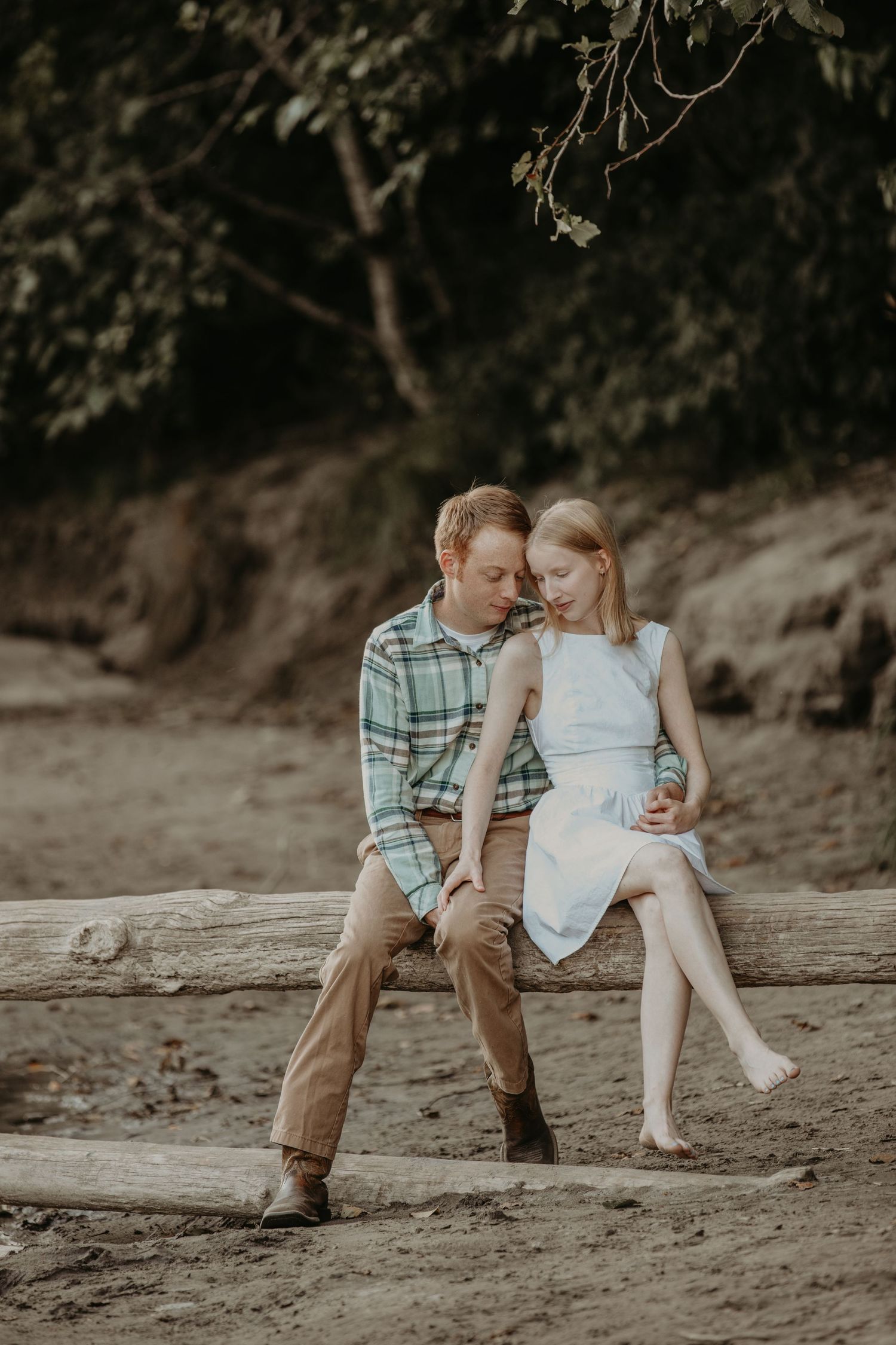 Young couple in casual attire sitting close together on a sandy beach during a romantic outdoor photoshoot.