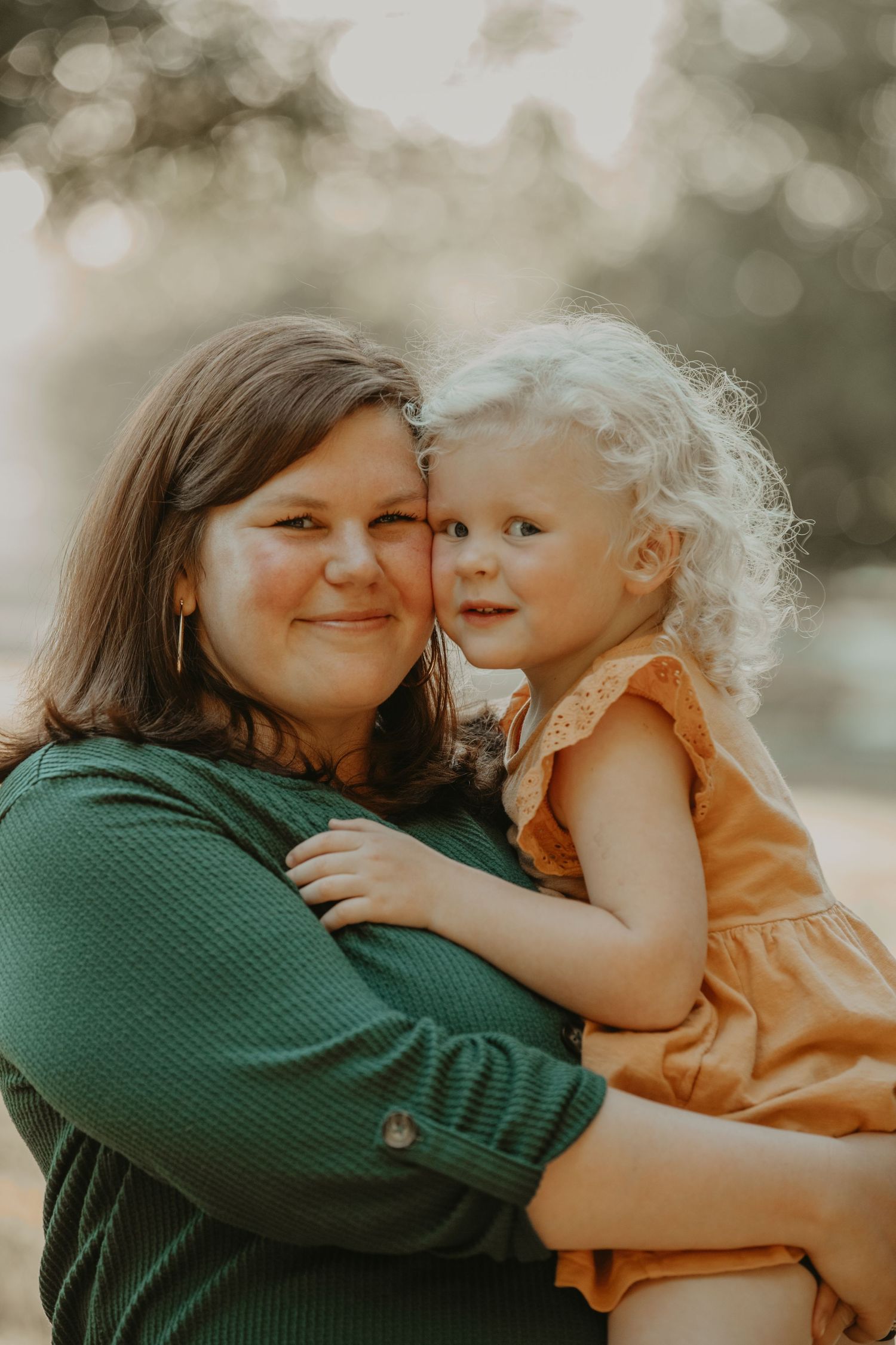 A tender moment captured outdoors between a woman in green and a young child in orange against a soft bokeh background.