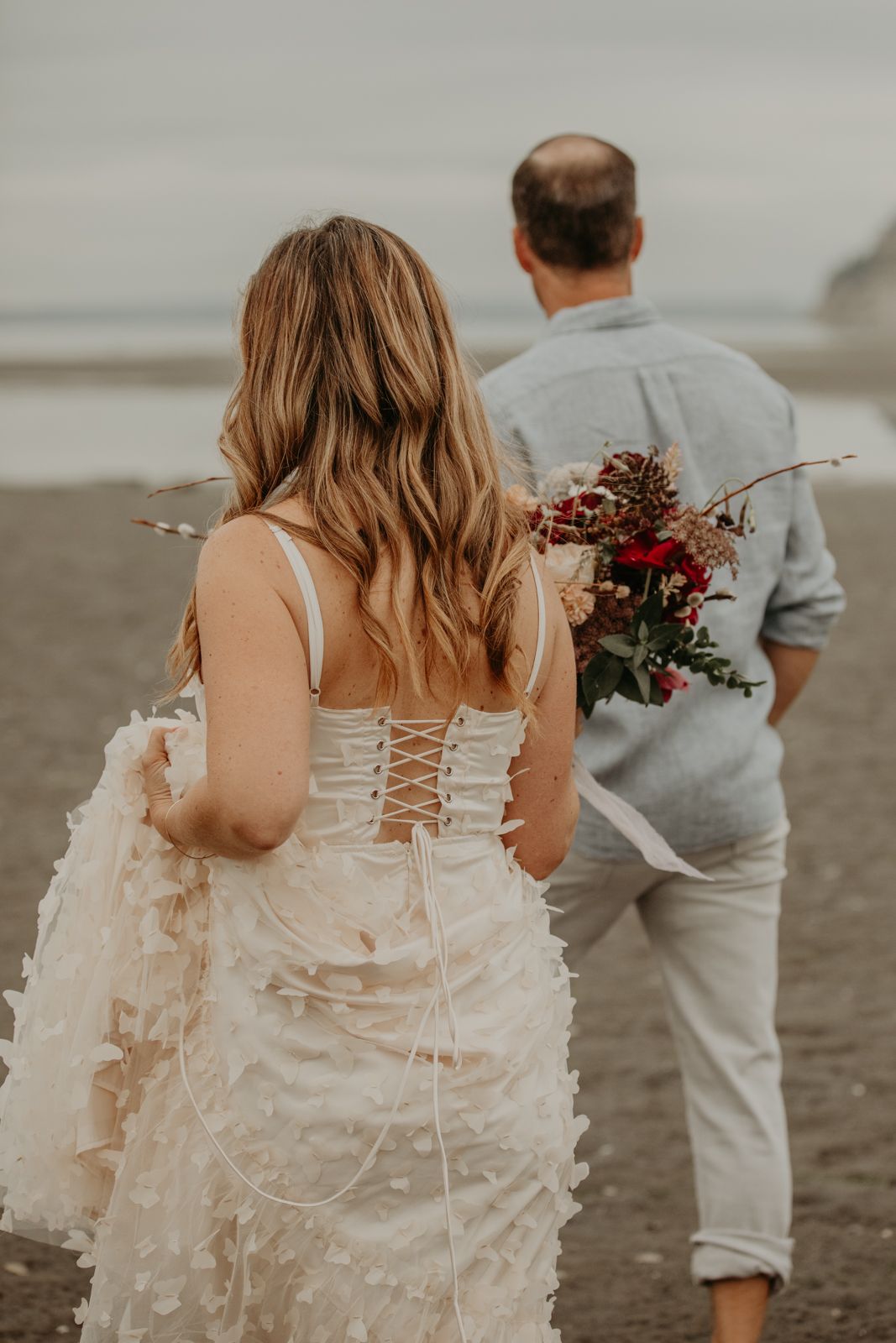 A romantic beach wedding scene showing the back of a bride in a lace-up dress with a floral bouquet walking on the sand.