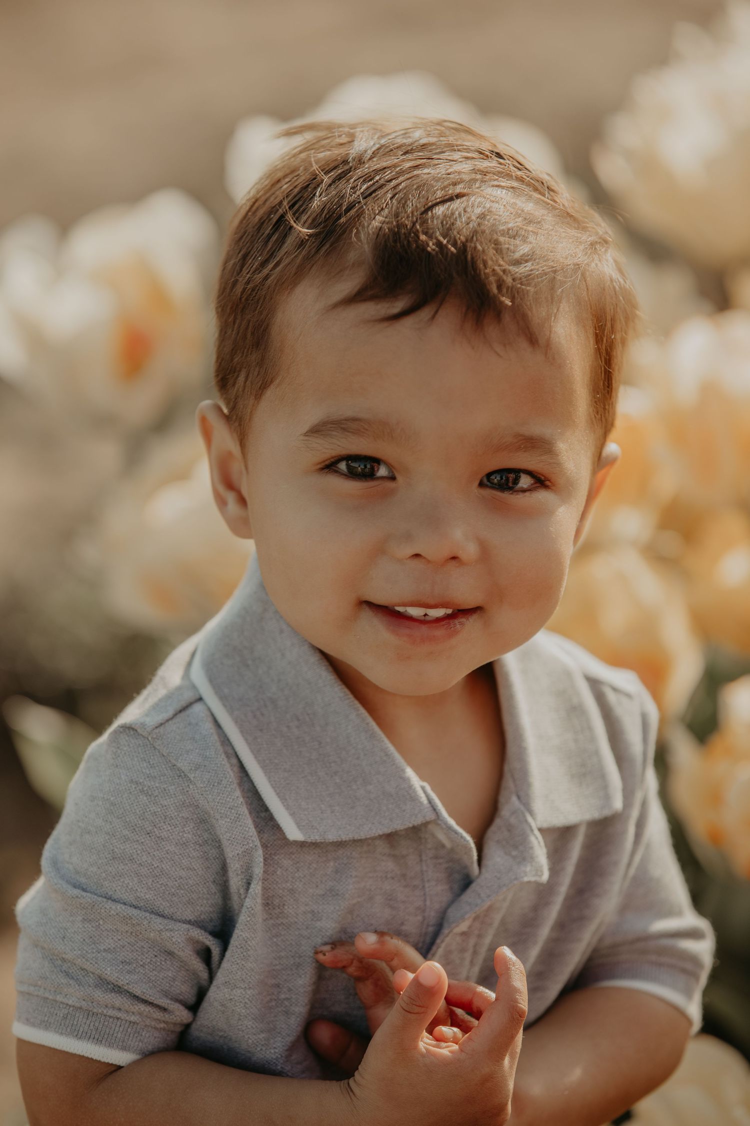 Portrait of a smiling toddler in a gray polo shirt outdoors with soft light and flowers in the background.