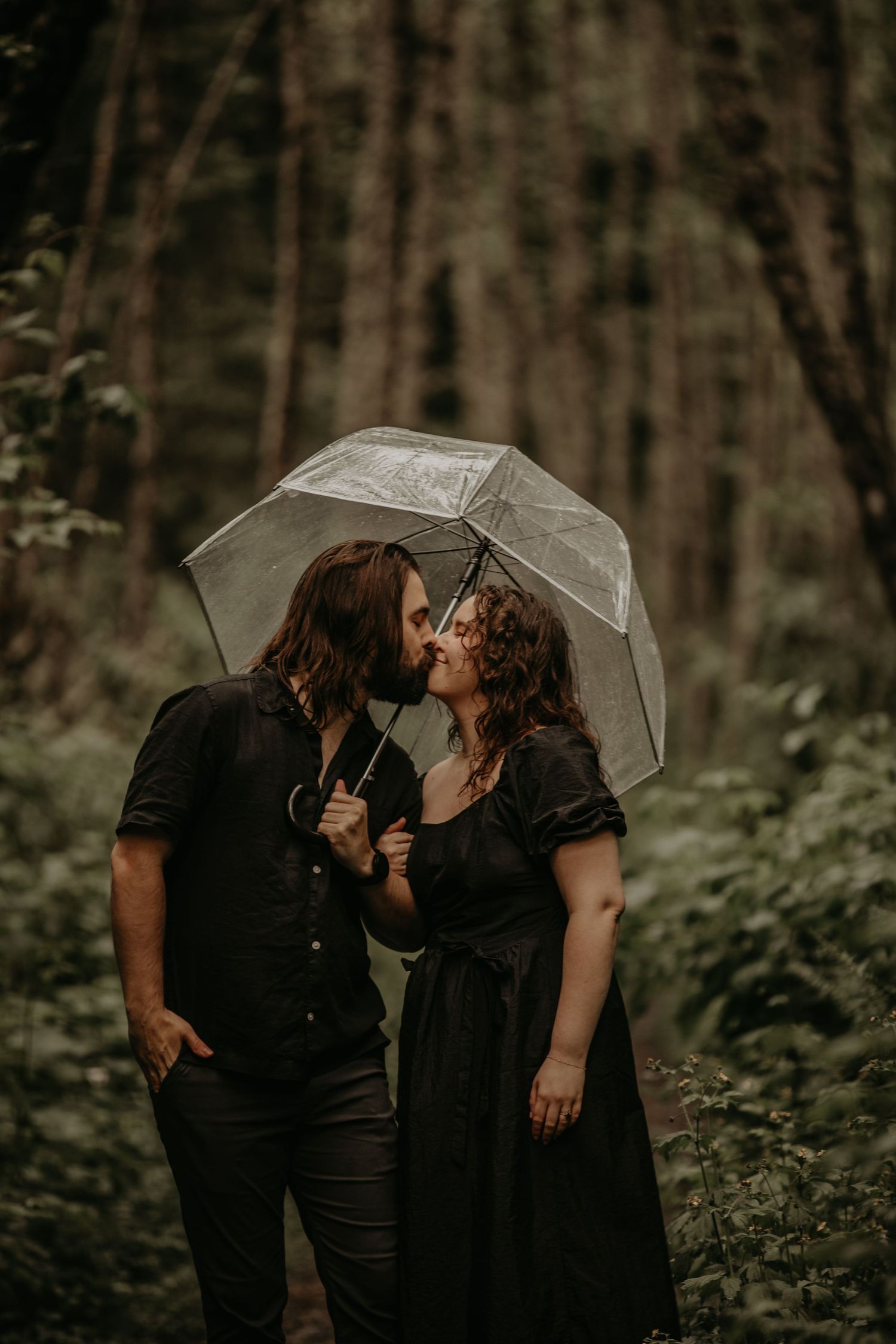 Intimate couple embracing under transparent umbrella in woodlands during romantic photoshoot.