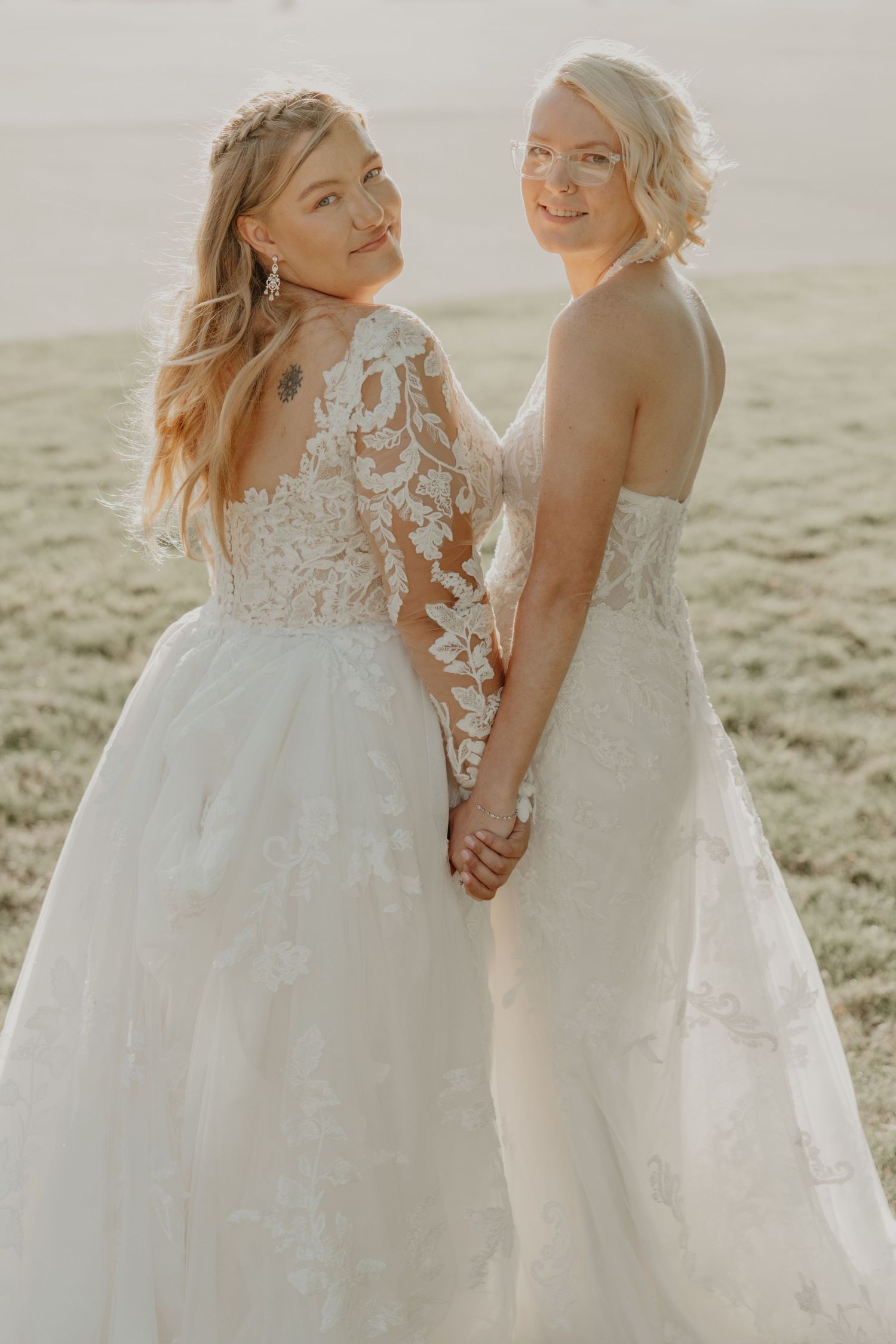 Two brides in elegant lace wedding gowns hold hands and smile while posing outdoors in a sunlit field.