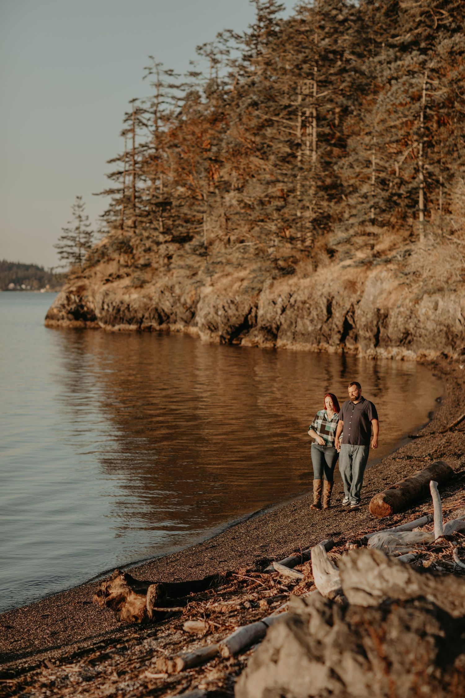 A couple stands on a rocky beach shoreline surrounded by tall evergreen trees and cliffs at golden hour.