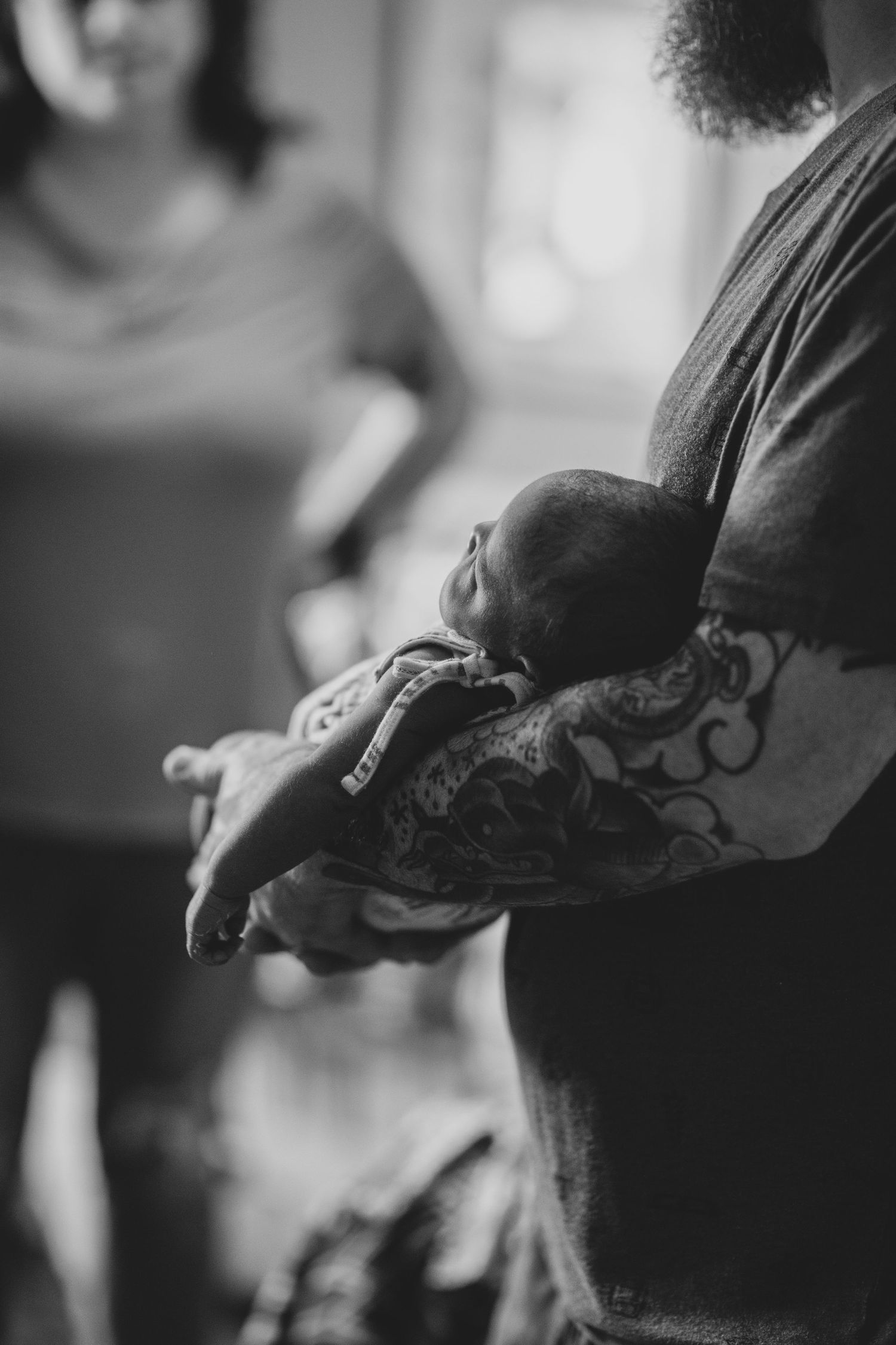 A tender black and white photograph shows tattooed arms gently cradling a sleeping newborn baby.