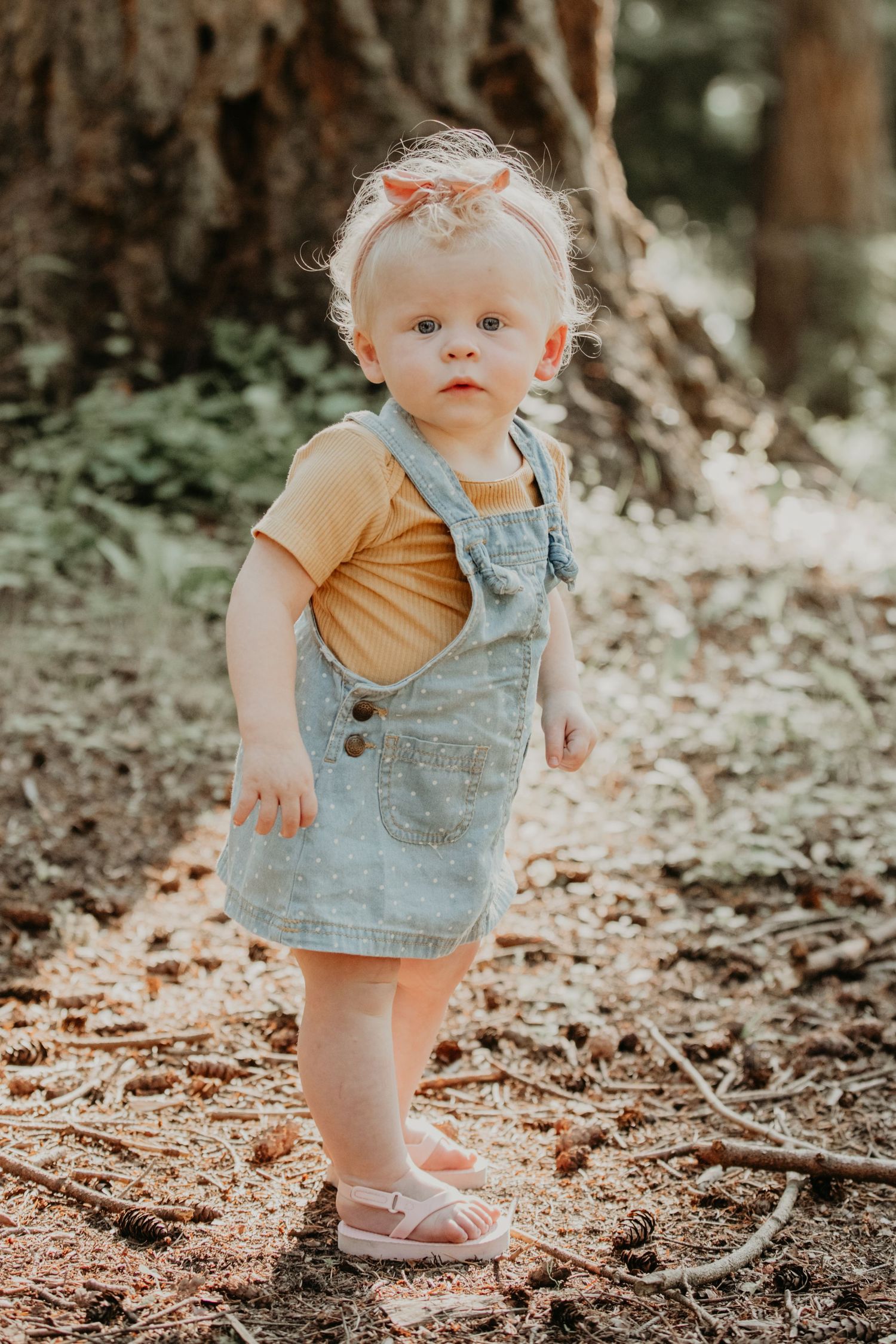Small child stands in a forest wearing denim overalls and tan shirt with matching bow on sunny afternoon.