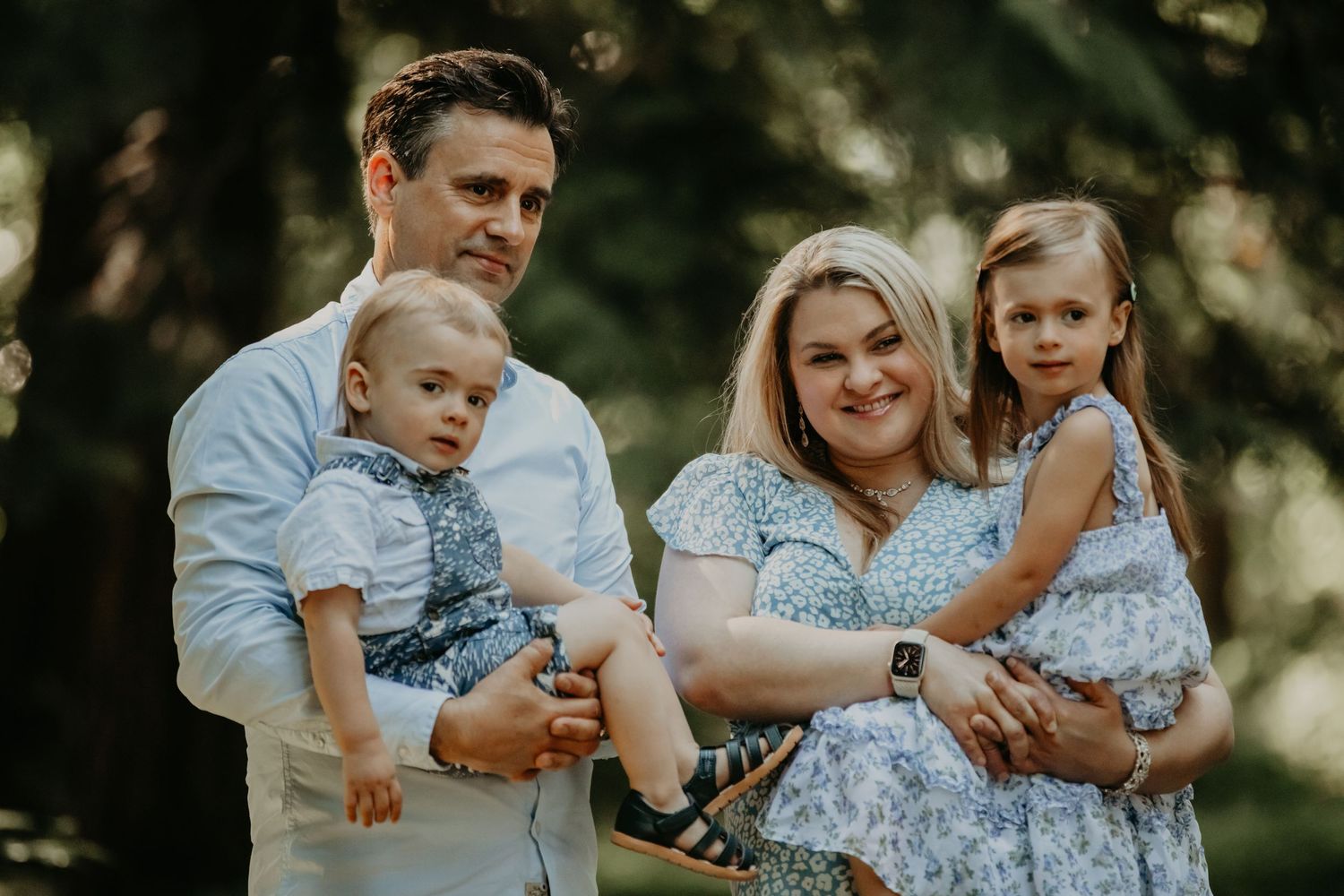 A family of four wearing white and blue outfits poses together outdoors in a sunlit wooded setting.