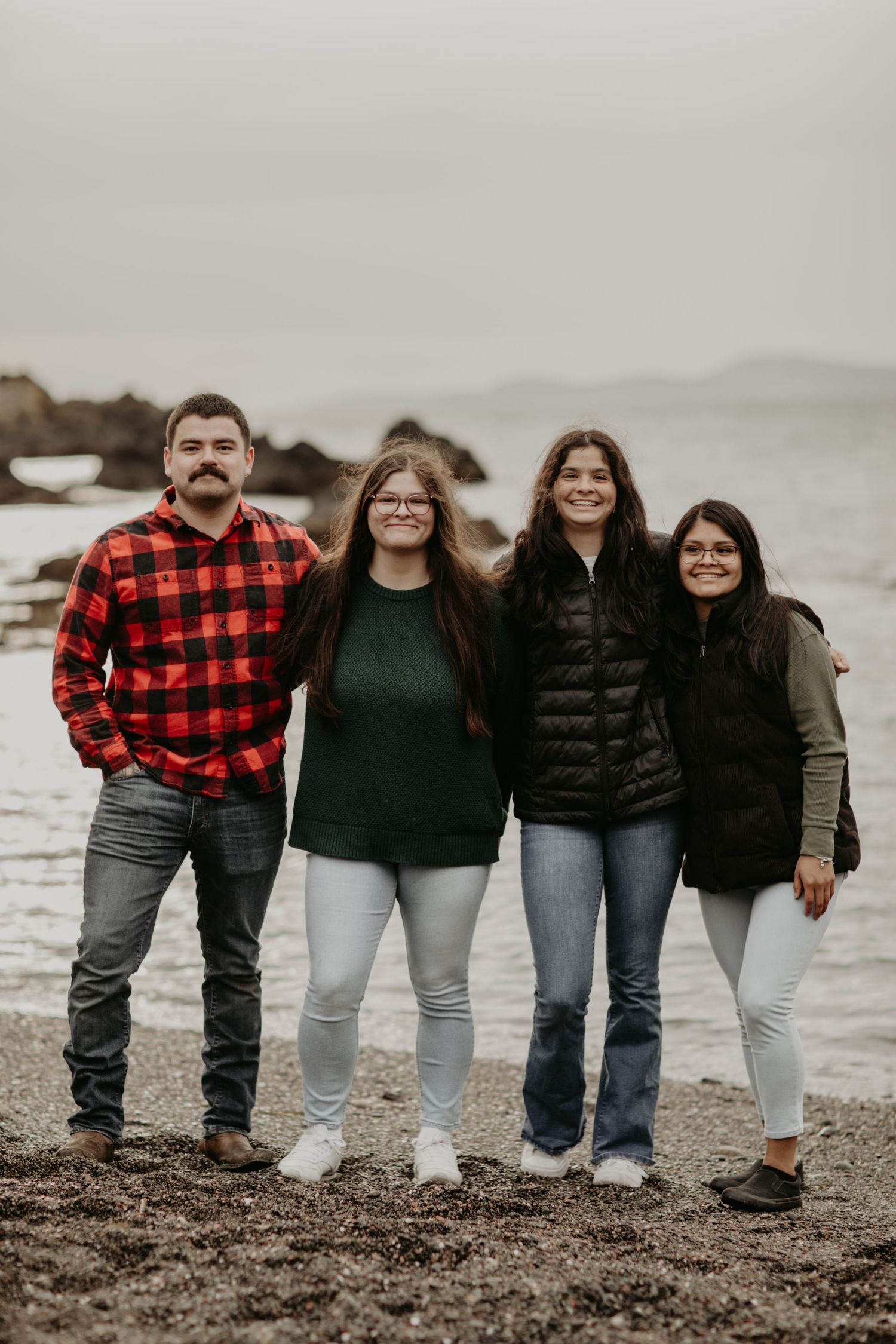 A group of four people stand together on a cloudy beach wearing casual attire including plaid and sweaters.