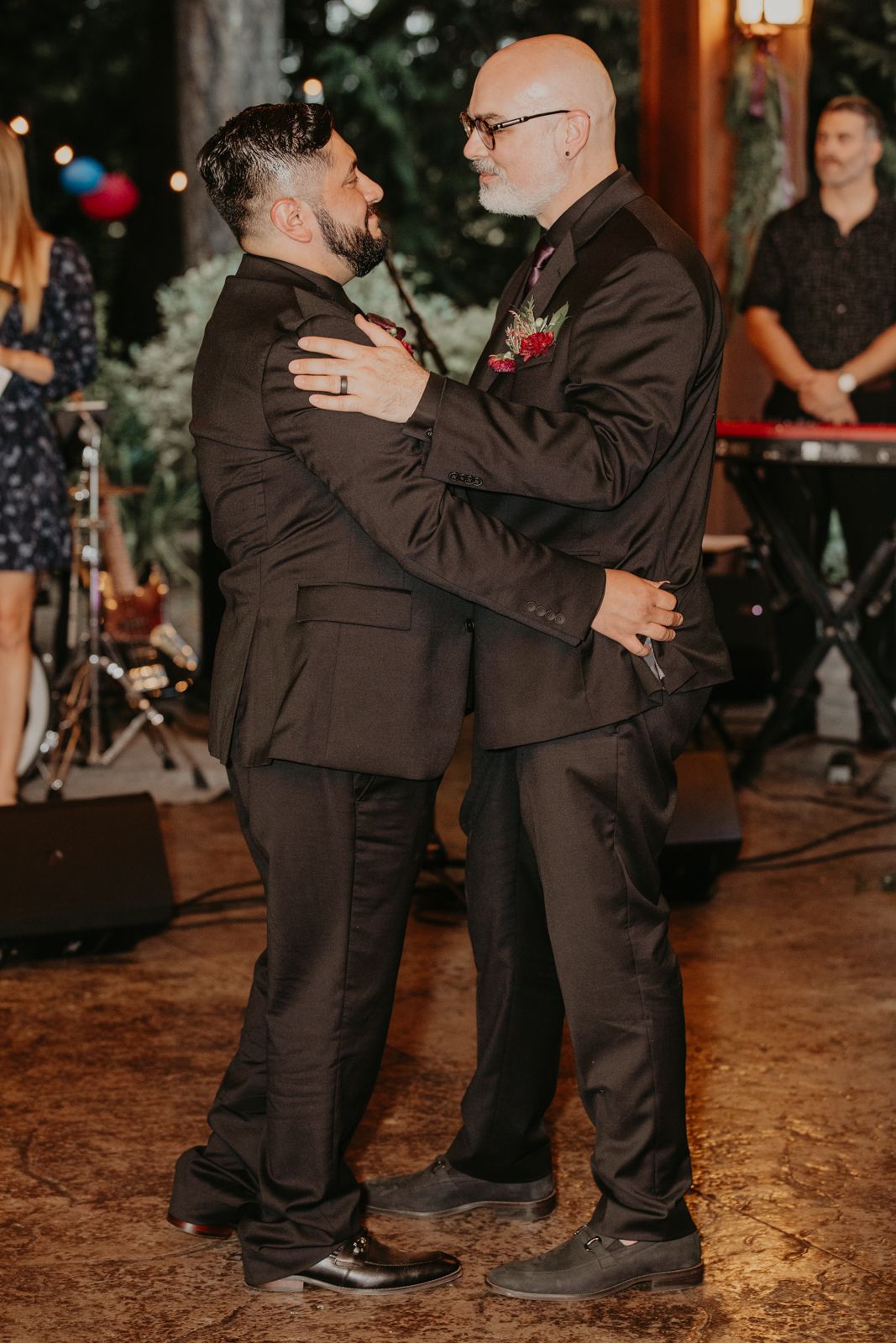 Two people in dark suits dance together on a rustic outdoor dance floor at an evening event.