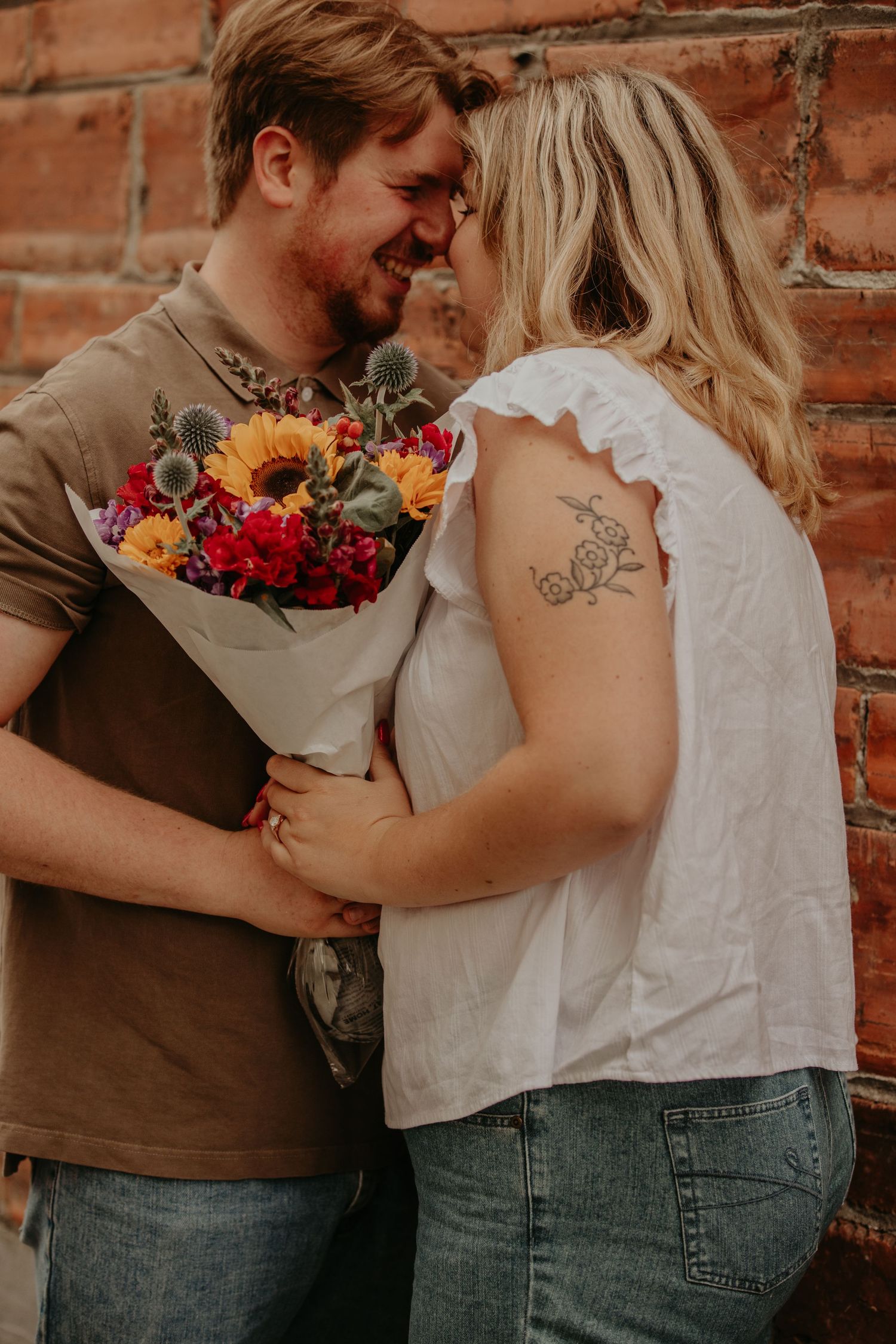 A romantic couple embraces while holding a vibrant bouquet against a rustic brick wall background.