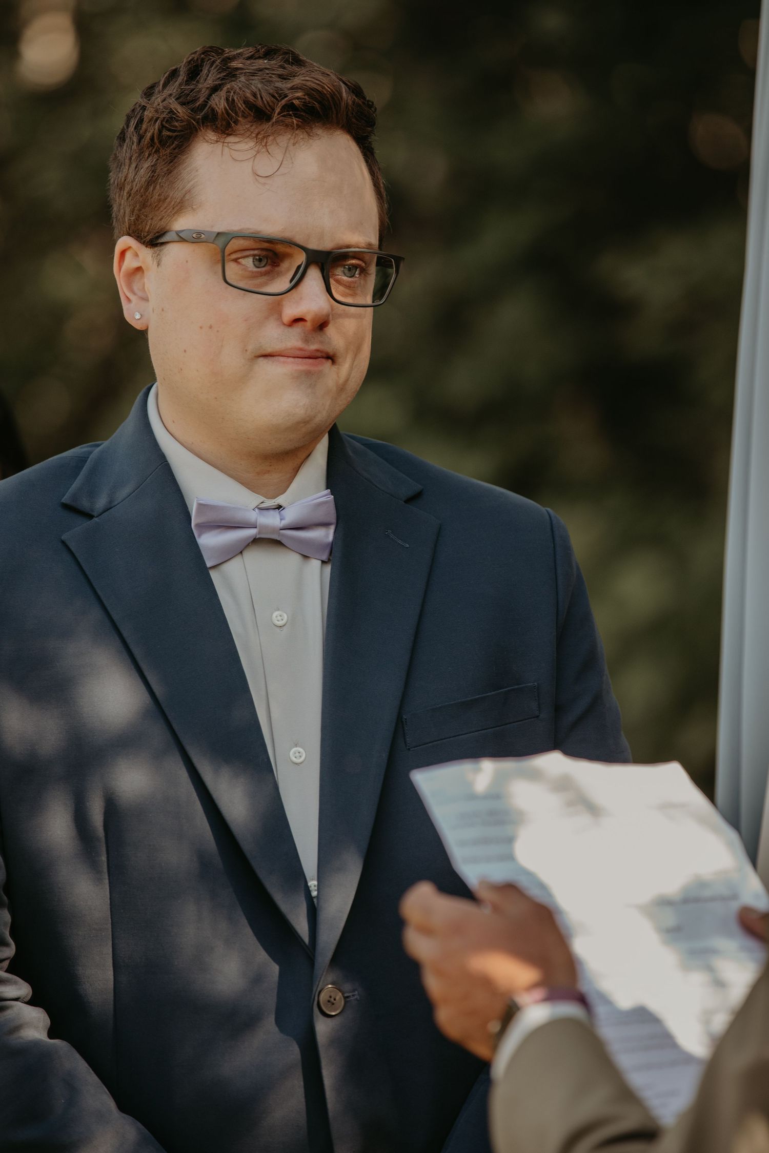 Person wearing glasses, navy suit and pink bow tie stands attentively during outdoor ceremony.