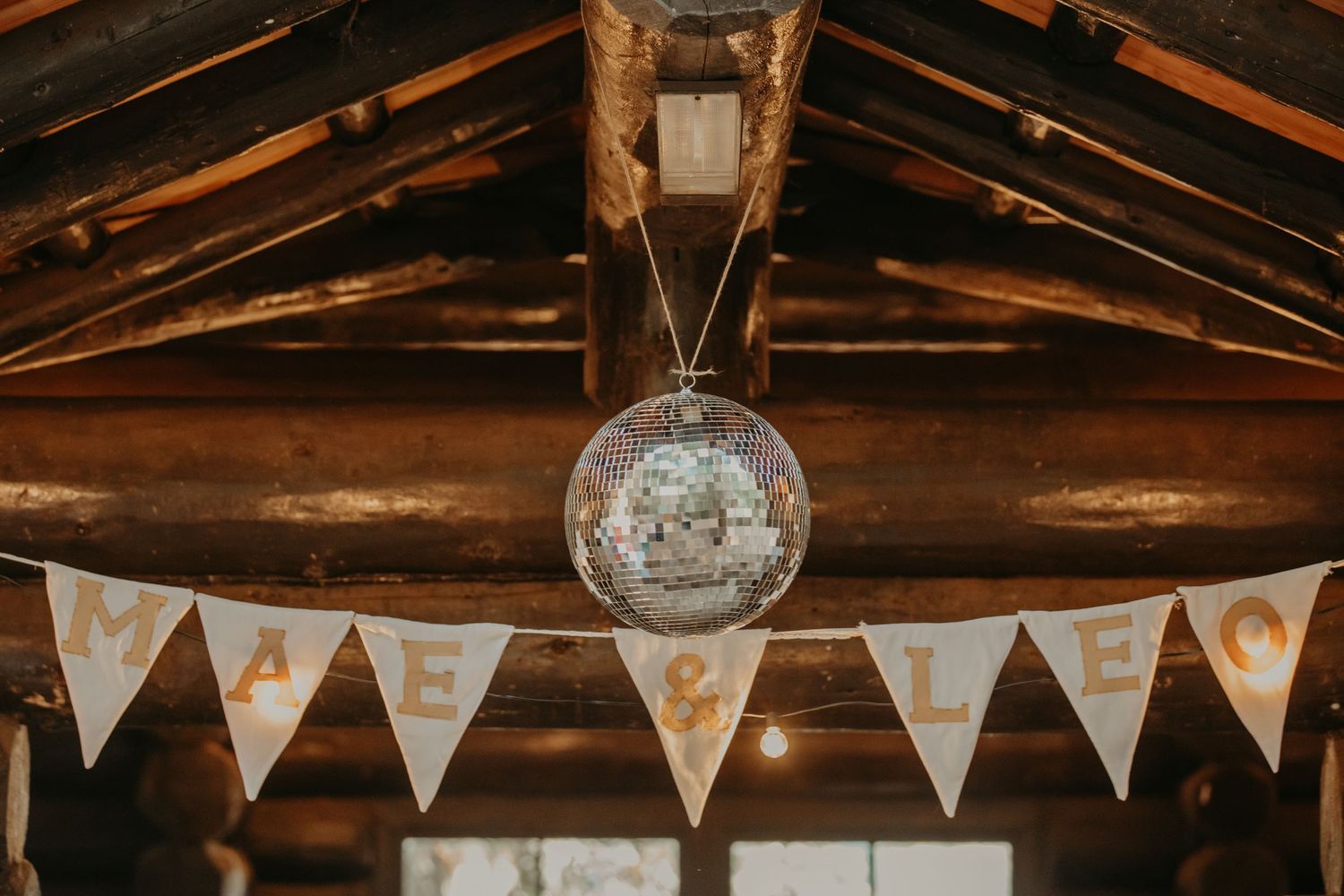 A disco ball hangs from rustic wooden rafters with white triangular wedding bunting flags below.