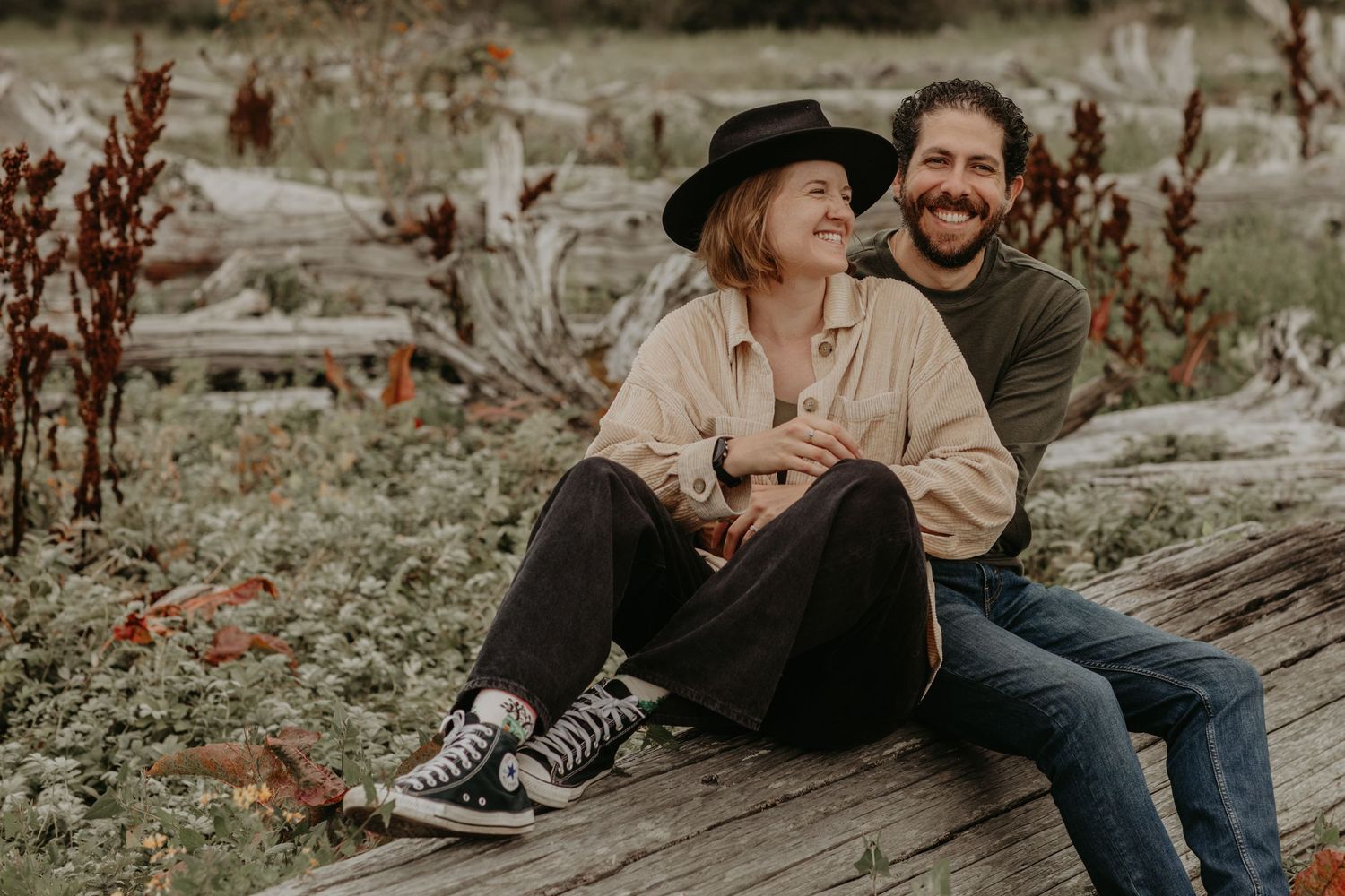 A couple sitting close together on rustic wooden boards surrounded by autumn foliage in a natural outdoor setting.