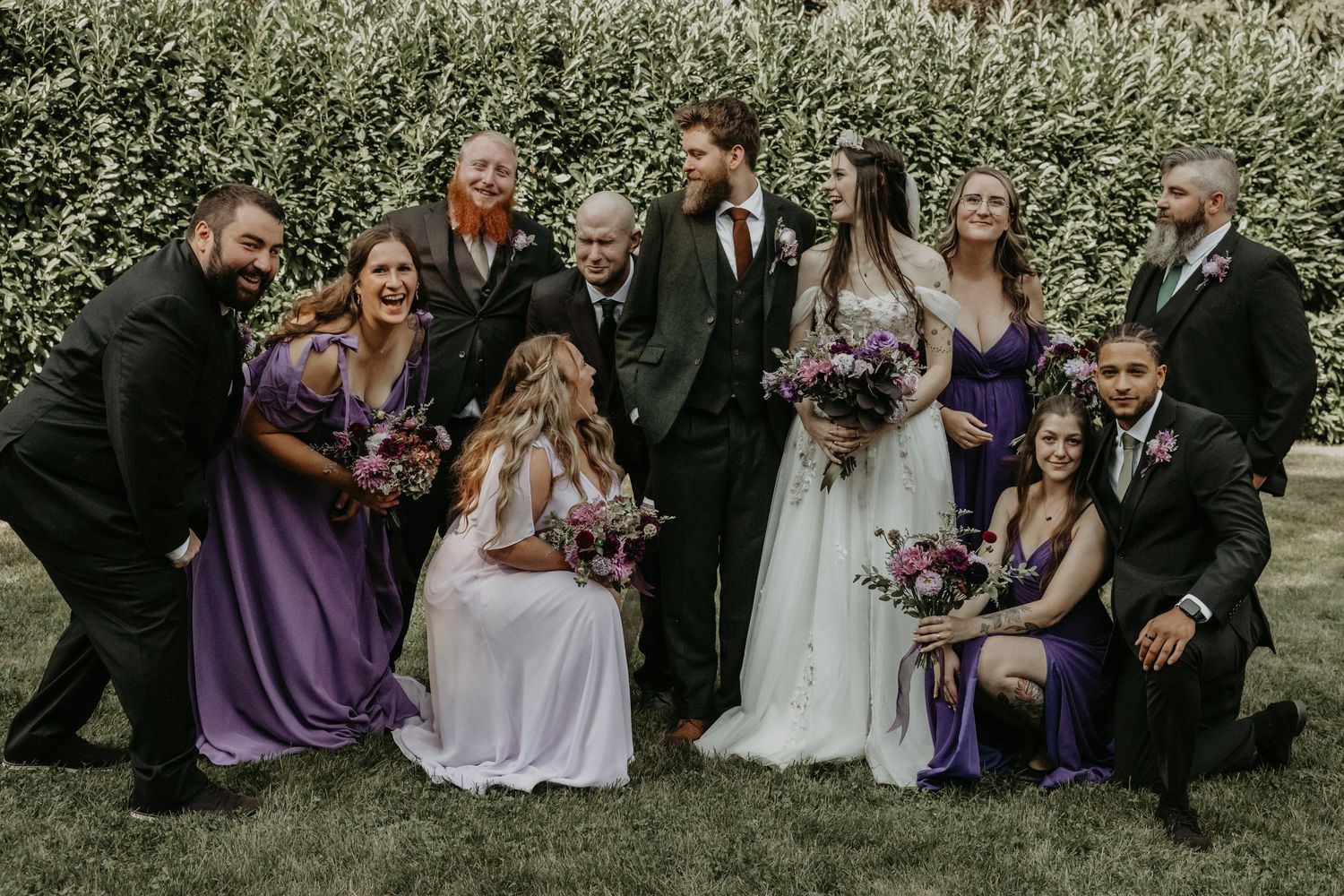 Wedding party in purple dresses and black suits poses together outdoors in a garden setting.