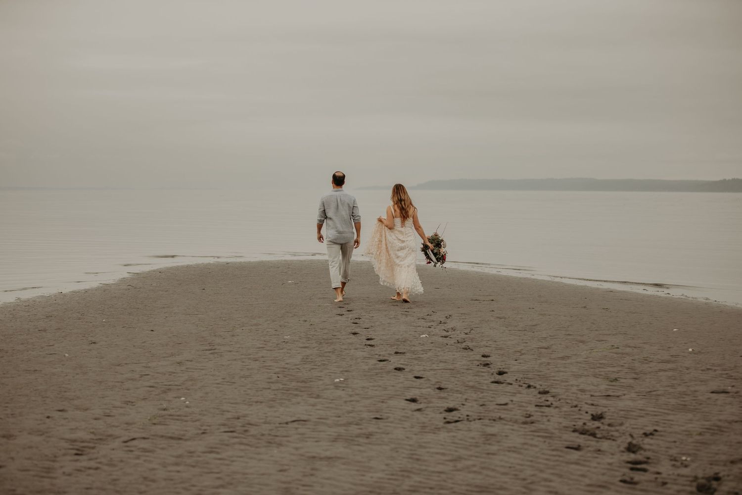 A couple in white wedding attire walks along a misty beach shore during an overcast day.