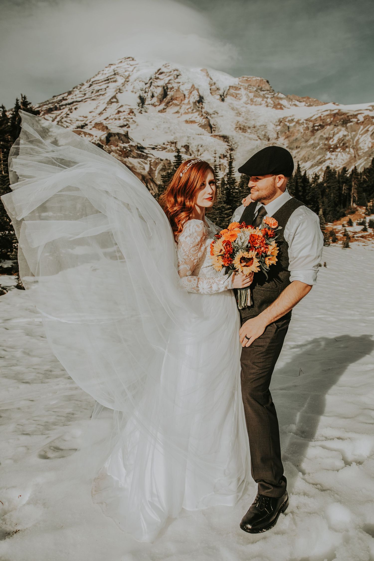 Wedding couple poses in snowy mountain landscape with flowing veil and sunflowers.