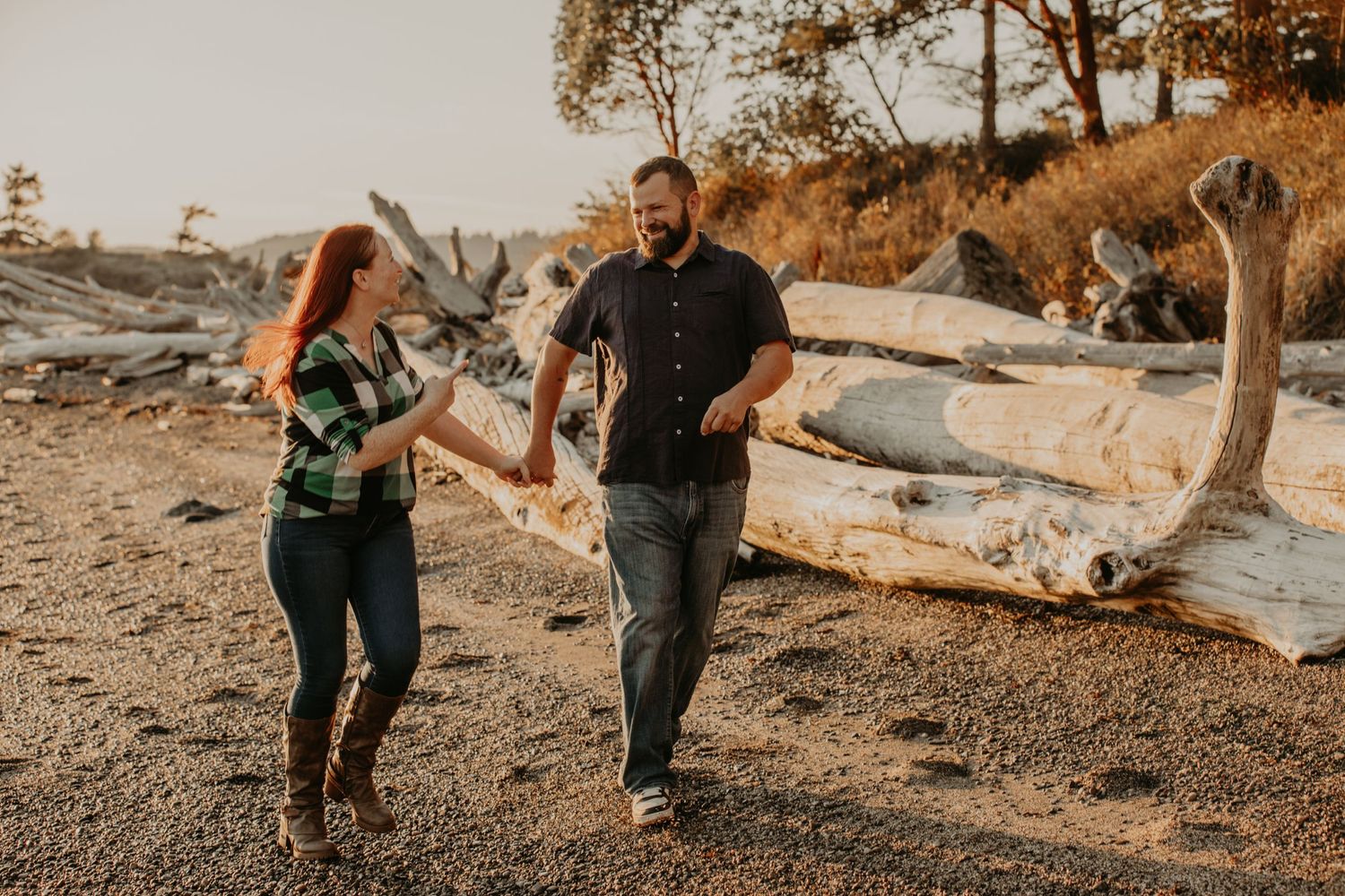 A couple walking and laughing together on a rocky beach with driftwood logs at sunset.