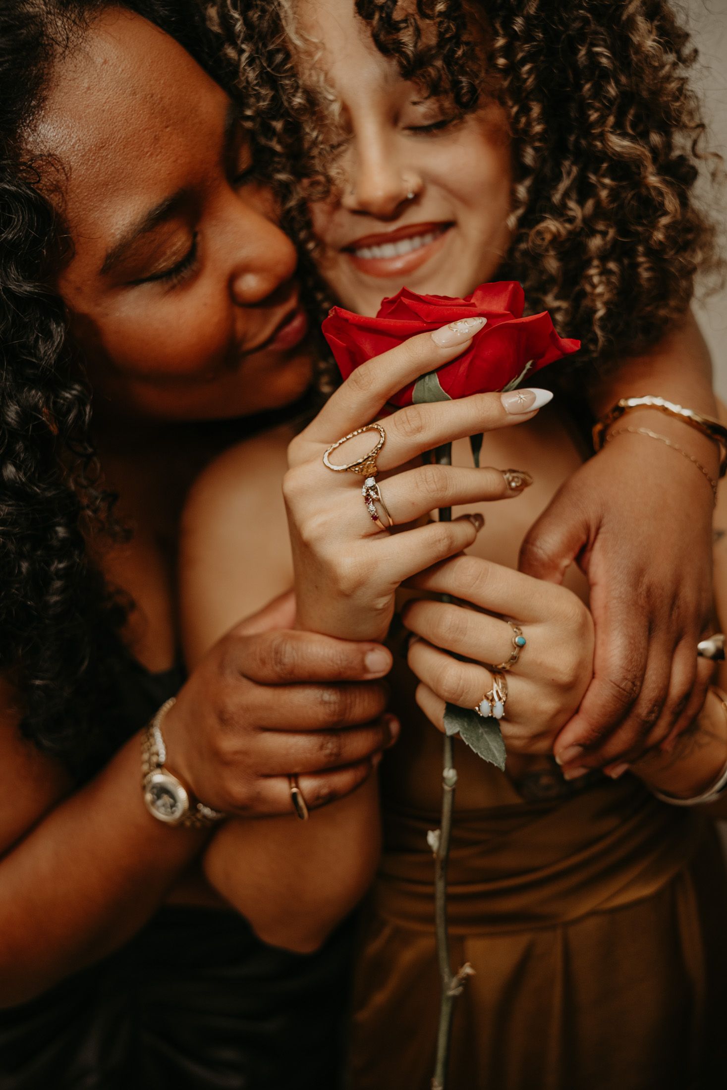 Two women with curly hair share a tender embrace while holding a red rose, adorned with rings and bracelets.