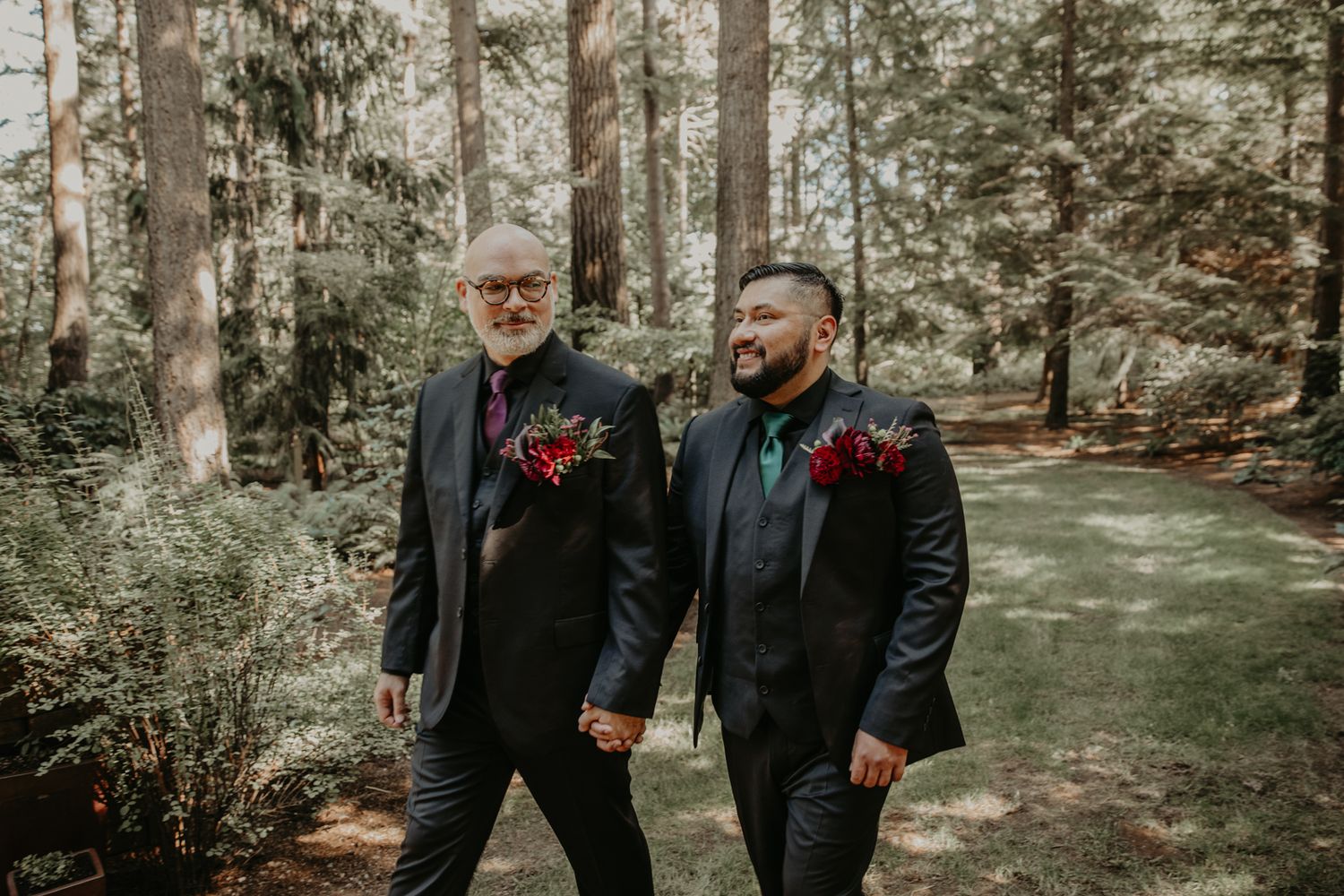 Two people in black suits with red boutonnieres hold hands while walking along a forest path.