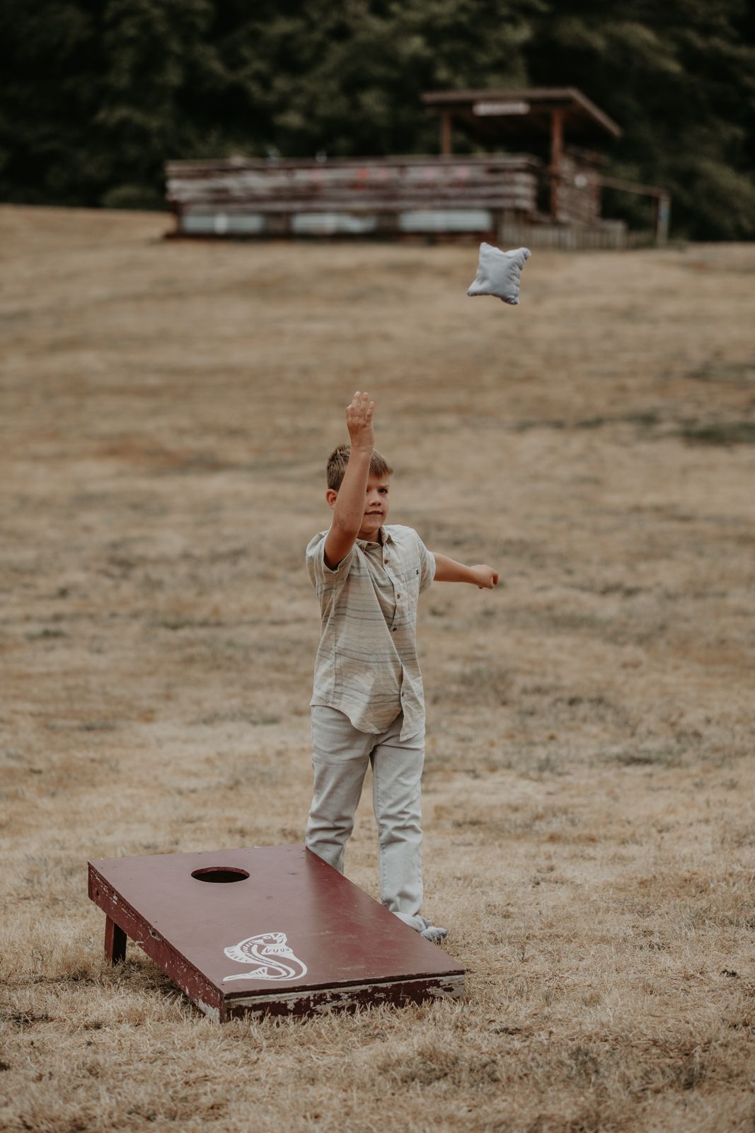 A person plays cornhole on a dry grass field, tossing bean bags toward wooden game boards.