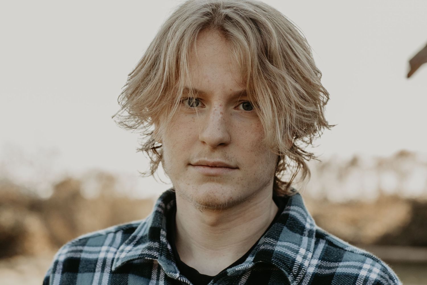 A person with wavy blonde hair wearing a blue plaid flannel shirt poses outdoors during golden hour.