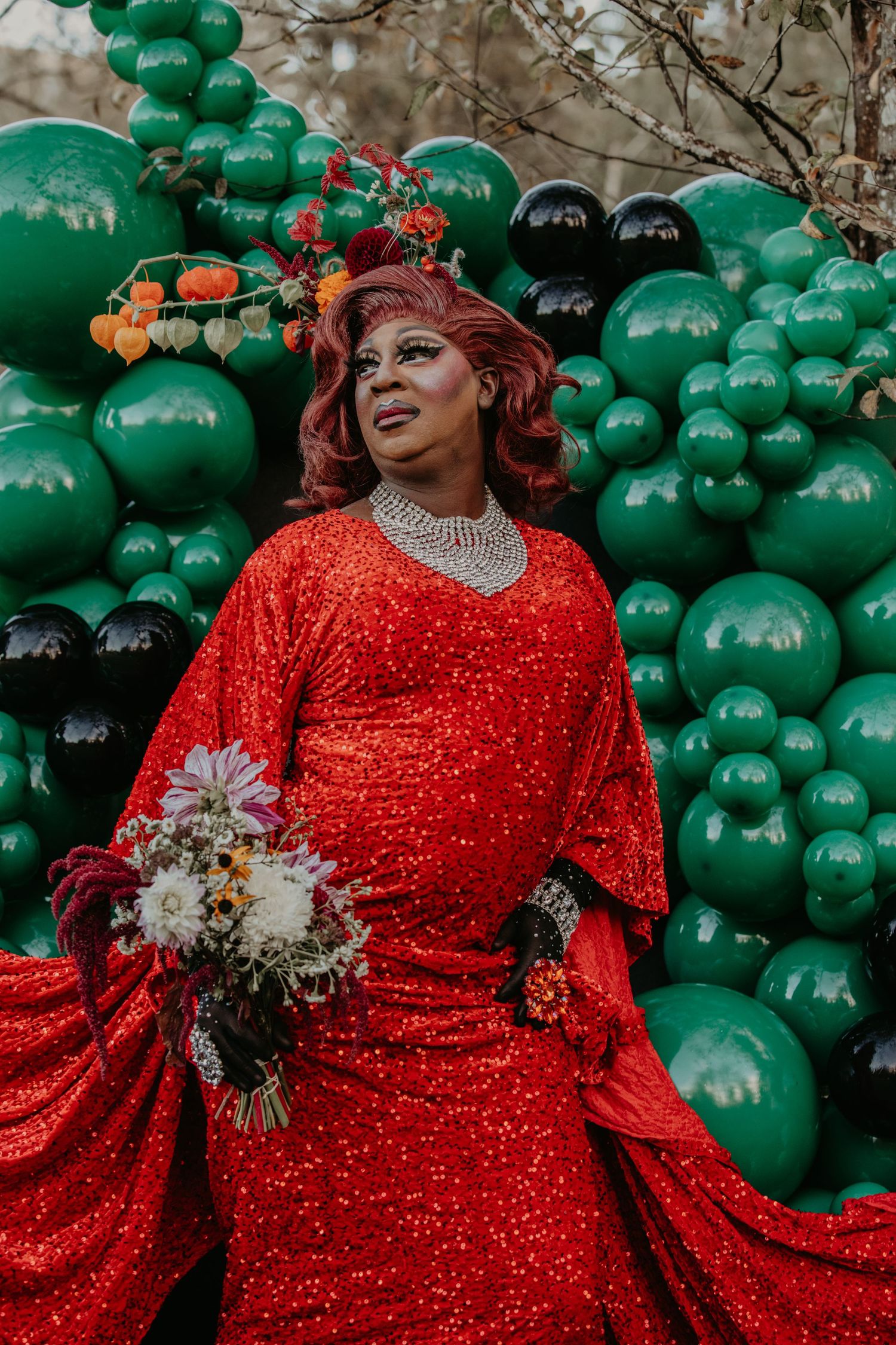 Glamorous figure in sparkly red gown poses against green and black balloon backdrop holding floral bouquet.
