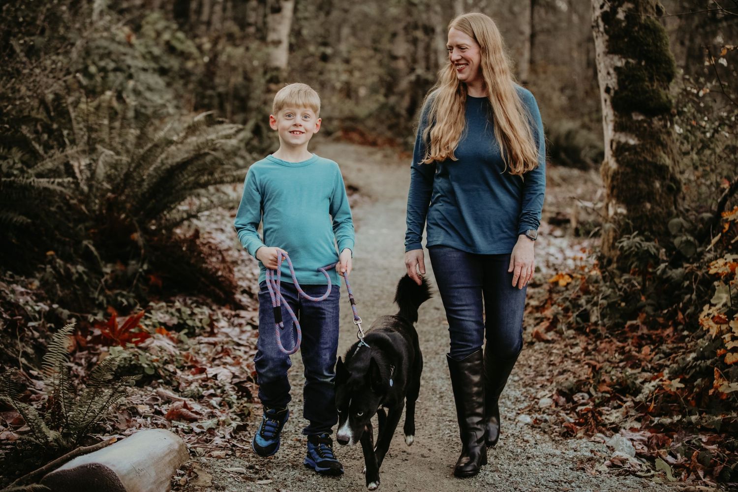 Two people enjoy an autumn walk with their black dog on a leaf-covered forest trail.