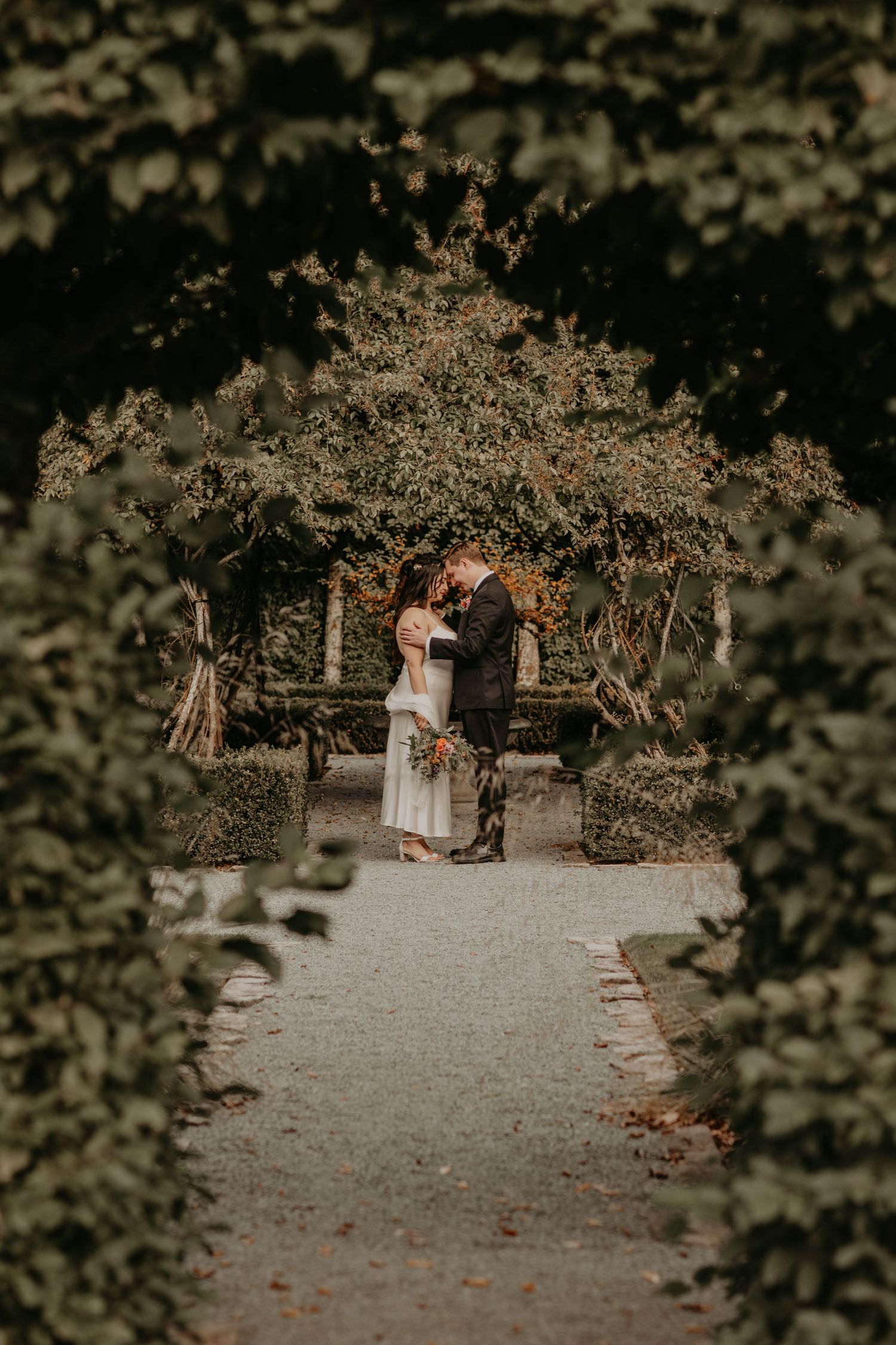 A couple shares a romantic moment on a garden path surrounded by lush greenery and fallen autumn leaves.