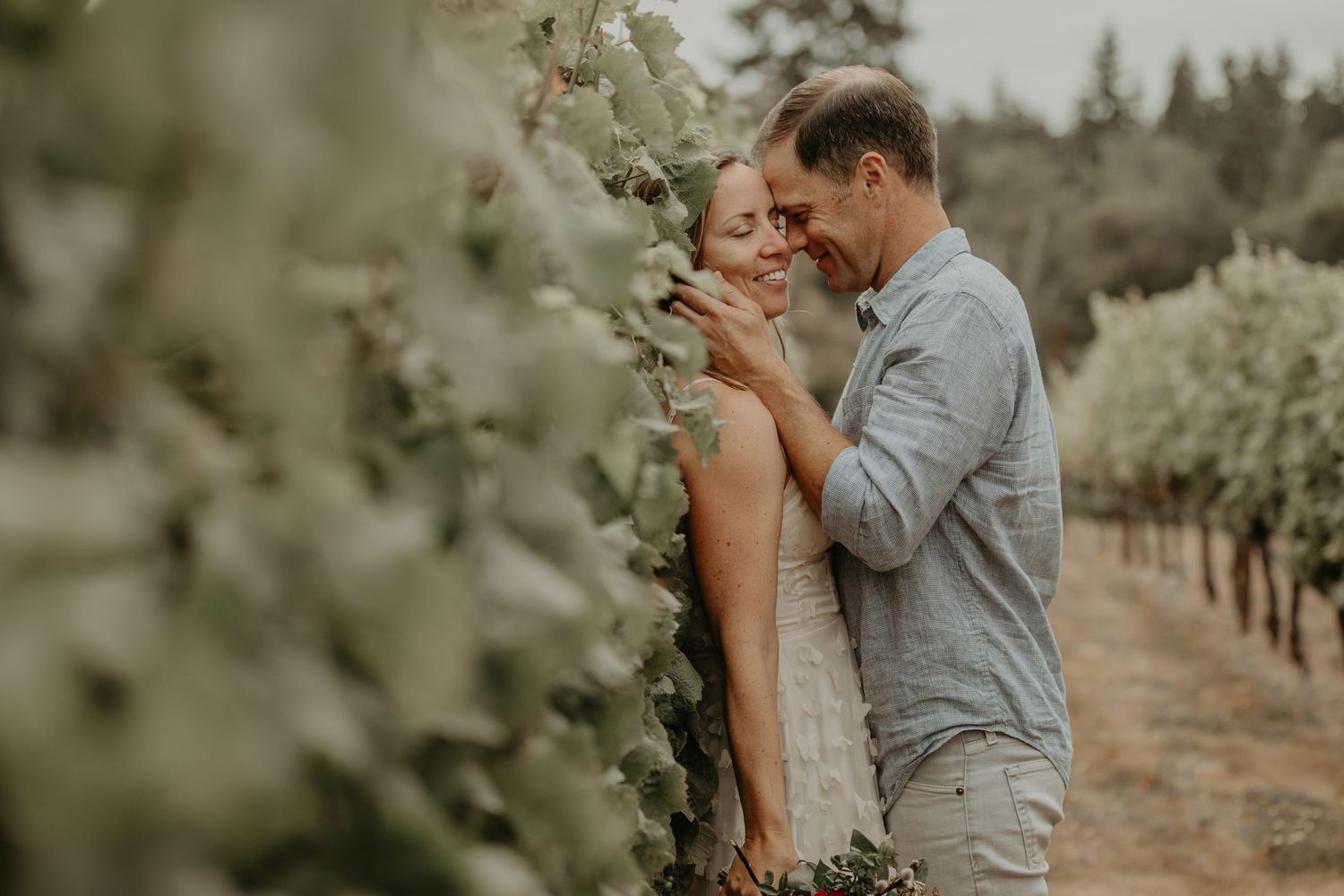 A couple shares an intimate moment in a rustic outdoor setting with muted green tones and natural lighting.