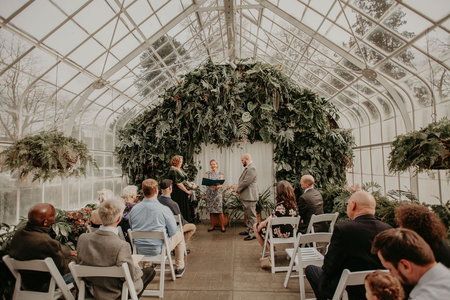Wedding ceremony takes place inside a greenhouse with floral arch and white chairs filled with guests.