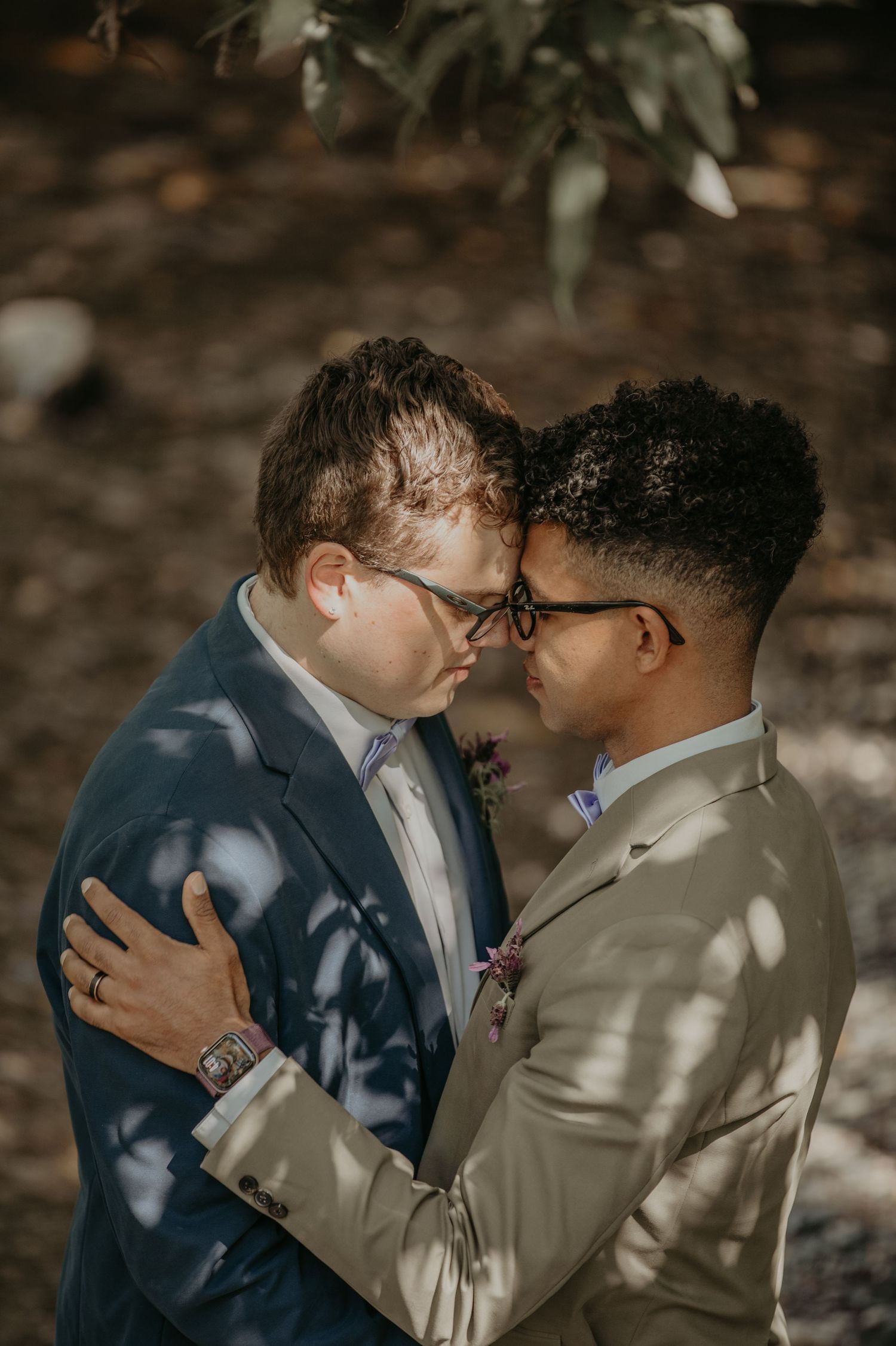 Two people in formal suits share a tender moment under dappled sunlight in a romantic outdoor setting.