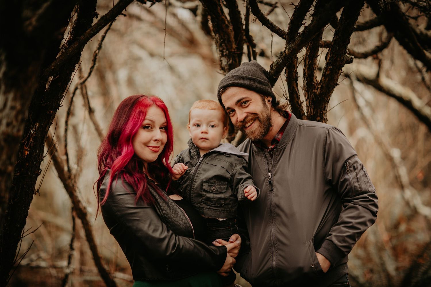 A family poses together in a moody autumn forest setting with bare branches creating a dramatic backdrop.