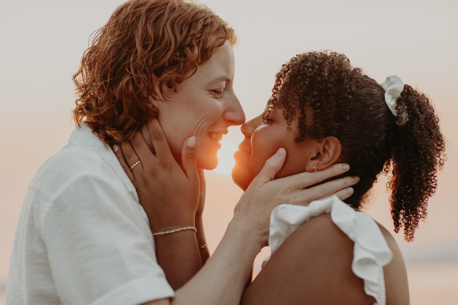 A mother and daughter share a tender nose-to-nose moment at sunset, smiling and holding each other's faces.