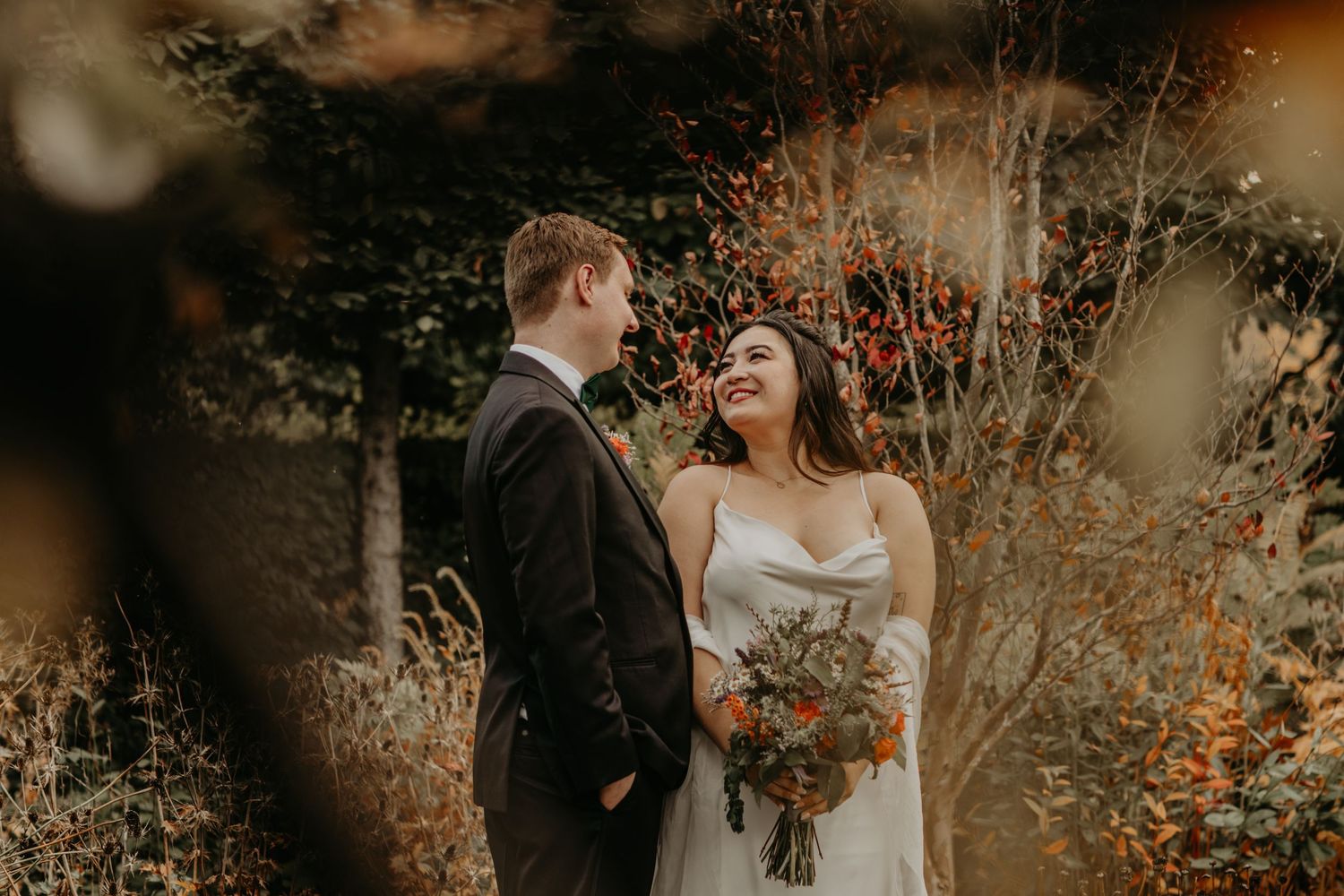 A candid moment of a bride and groom sharing a laugh together in a warm autumn setting with golden dried foliage.
